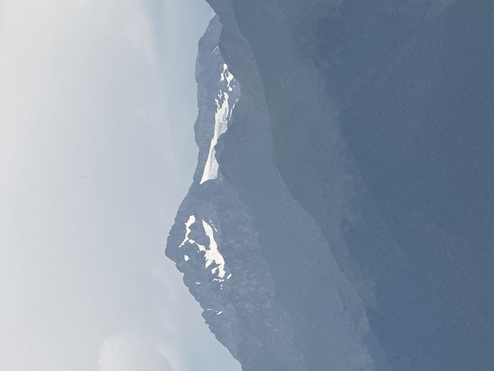 An image captures a view of multiple mountain ranges layered against a pale, overcast sky. The foreground consists of dark, blue-gray mountains that recede into the distance. The midground shows a lighter, bluish-gray mountain range, also appearing somewhat hazy. The background features the tallest mountain, with a rugged, dark gray summit. Patches of snow are visible on the upper slopes of this prominent peak. The overall atmosphere is muted and somewhat soft due to the cloudy conditions. No people, buildings, or specific urban features are discernible in the image.