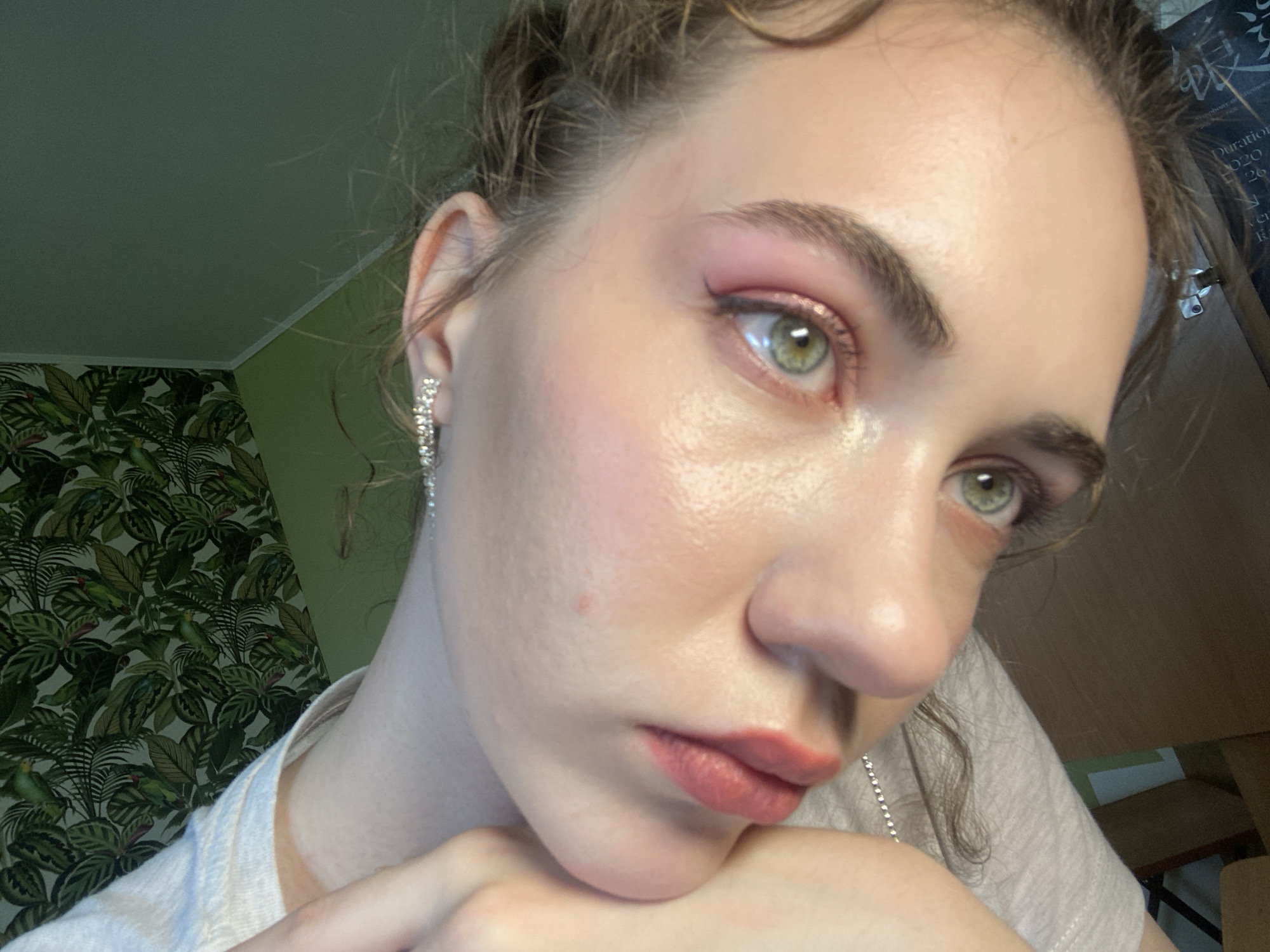 The image is a close-up, low-angle shot of a young woman's face. She has fair skin, light brown hair styled in loose curls, and green eyes. She is wearing pink eyeshadow and a light pink lipstick. She is also wearing a sparkly earring in her left ear. Her hands are resting near her chin, and she is wearing a light-colored t-shirt. In the background, there is a wall with tropical-themed wallpaper featuring green leaves and parrots. To the right of the woman, a table and some boxes are visible. The overall lighting suggests an indoor setting, likely during the day. There is no specific information or visual cue within the image to identify the location as Zavodoukovsk, Russia.