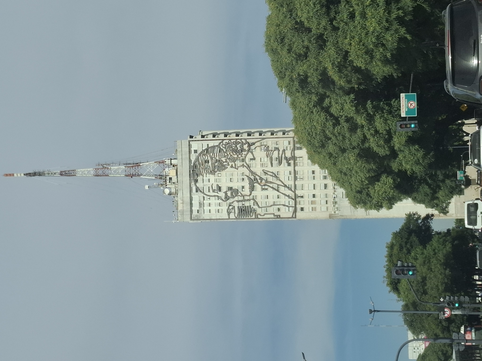 The image depicts a street scene in Buenos Aires, Argentina. A tall building with a mural of a human face, possibly a historical figure, is the focal point. A transmission tower is attached to the building's side. Dense green trees partially obscure the building and line the street. Streetlights and traffic signals are visible, with at least one displaying a green light. Vehicles, including a black Jeep and a white van, are on the road. A street sign indicates "Atencion". The sky is blue with some cloud cover.