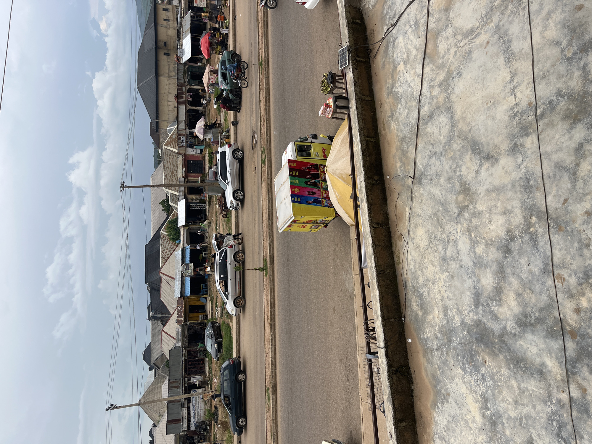 The image depicts a street scene in Rafin Par, Nigeria, on an overcast day. The street is lined with small shops and buildings with corrugated metal roofs. Several cars, including a silver SUV and a dark sedan, are parked or driving on the road. A yellow three-wheeled vehicle, decorated with colorful images of people, is parked on the side of the road. In the foreground, a concrete ledge overlooks the street, with a solar panel and wires visible on the ledge. The sky is heavily clouded, suggesting potential rain. The general atmosphere appears to be that of a busy commercial area with a mix of vehicles and pedestrians.
