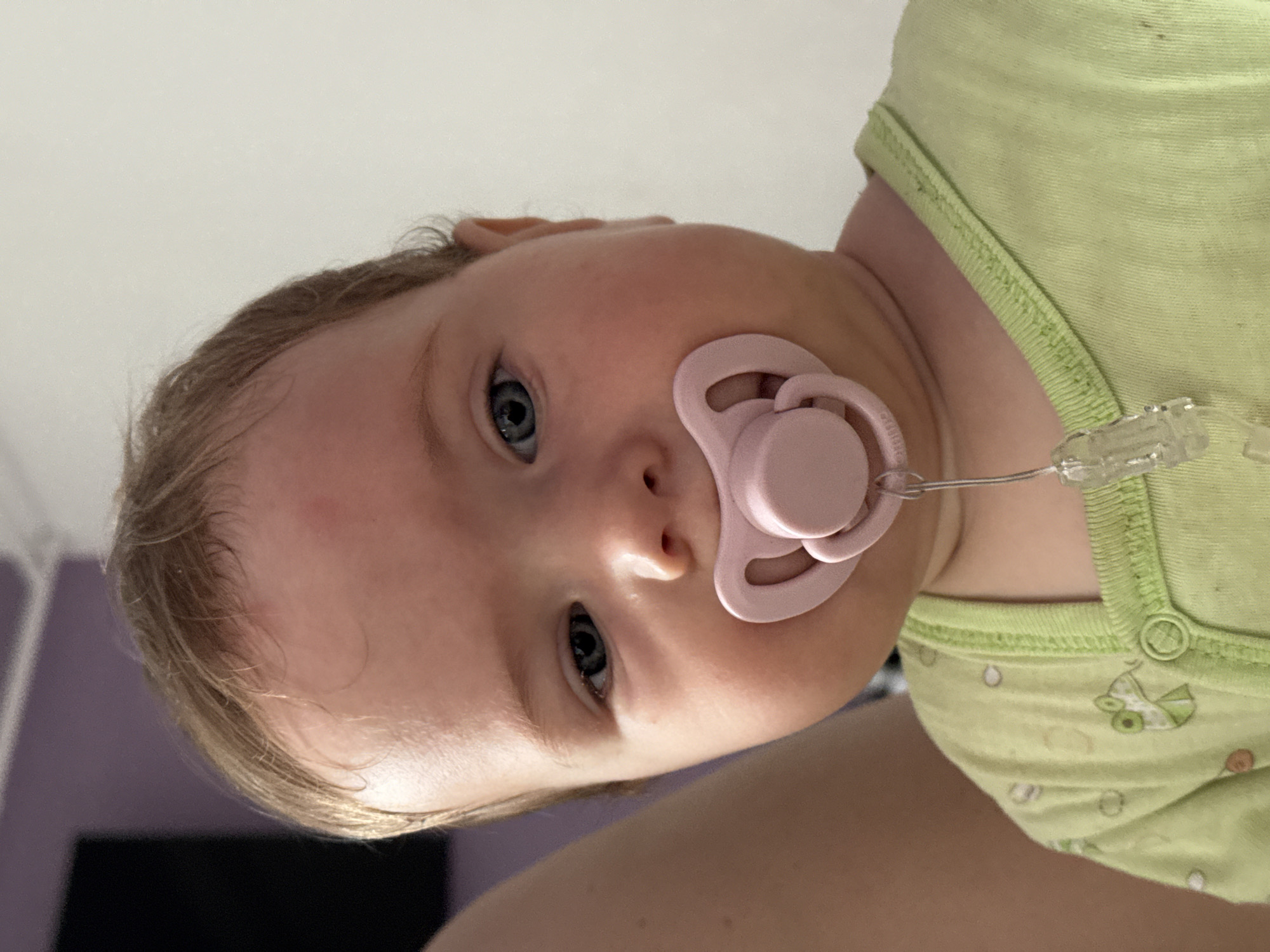 A close-up, top-down view captures a baby lying down and looking directly at the camera. The baby has fair skin, light-colored hair, and wide, bright blue eyes. A pink pacifier is in the baby's mouth, attached via a clear plastic clip to its clothing. The baby is wearing a light green short-sleeved bodysuit or onesie, adorned with a subtle pattern that includes what appears to be a small pram or stroller graphic.
The background is predominantly a plain, light color, suggesting an indoor setting. A darker, possibly purple or grey, area is visible on the left side of the frame, behind the baby's head. The baby's expression is neutral and observant. The image is taken in Rusalevka, Russia. No specific time of day or weather conditions can be determined from the indoor context. There is no visible text within the image.