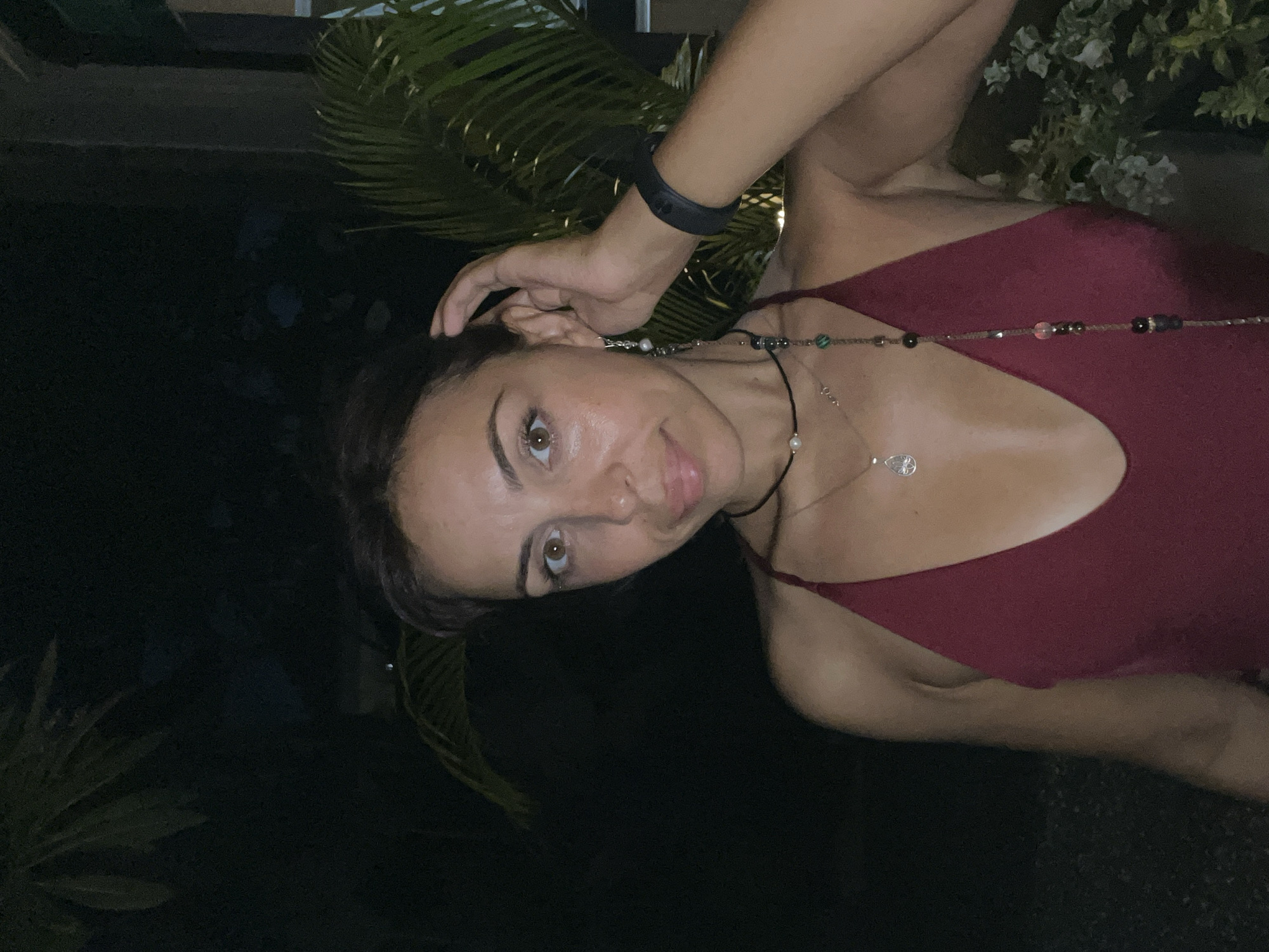 The image shows a close-up portrait of a young woman, captured in a chest-up frame. Her head is slightly tilted, and she is looking directly at the camera with a calm, observant expression. Her dark hair appears pulled back, and she has visible brown eyes and full lips, slightly parted.
She is wearing a dark red or maroon camisole or tank top. Her left arm is raised, with her fingers gently touching the side of her head or hair, creating a relaxed pose. On her left wrist, she wears a black wristband, possibly a fitness tracker or watch. She is adorned with two necklaces: a longer, colorful beaded necklace featuring various shades including green, pink, silver, and black, and a shorter, delicate silver chain necklace with a small pearl and a heart-shaped or teardrop pendant, resting near her collarbone. A small earring is visible on her left ear.
The background is very dark, indicating it is nighttime. Lush green tropical foliage, resembling palm fronds and other leafy plants, is subtly visible in the upper right and along the top and bottom edges of the frame. This suggests an outdoor or semi-outdoor setting, such as a garden, patio, or balcony, consistent with the Kuta Selatan, Indonesia context. The woman is illuminated by what appears to be a direct light source, highlighting her features against the dark surroundings. There is no visible text in the image.