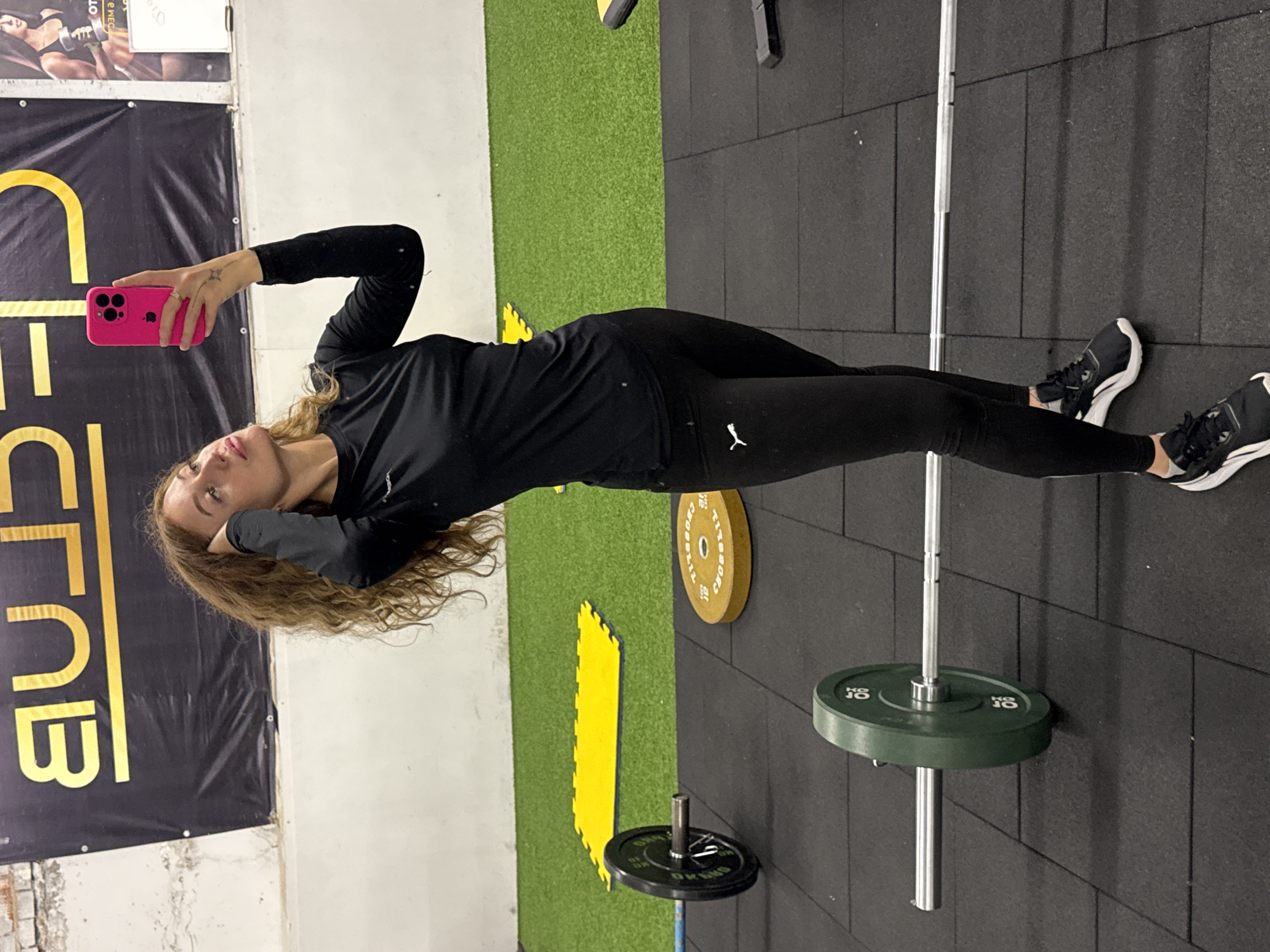 A woman with long, curly blonde hair is taking a selfie in a gym. She is wearing a long-sleeved black shirt and black leggings, and she is holding a pink cell phone in her left hand. She is standing on a black rubber floor with white grout lines. To her left is a banner with the word "CLUB" printed vertically in gold letters on a black background. To her right, there is a barbell with green weight plates and a yellow weight plate. In the background, there is a section of green turf. The location is potentially a gym in the city of Chaikovsky, Russia.