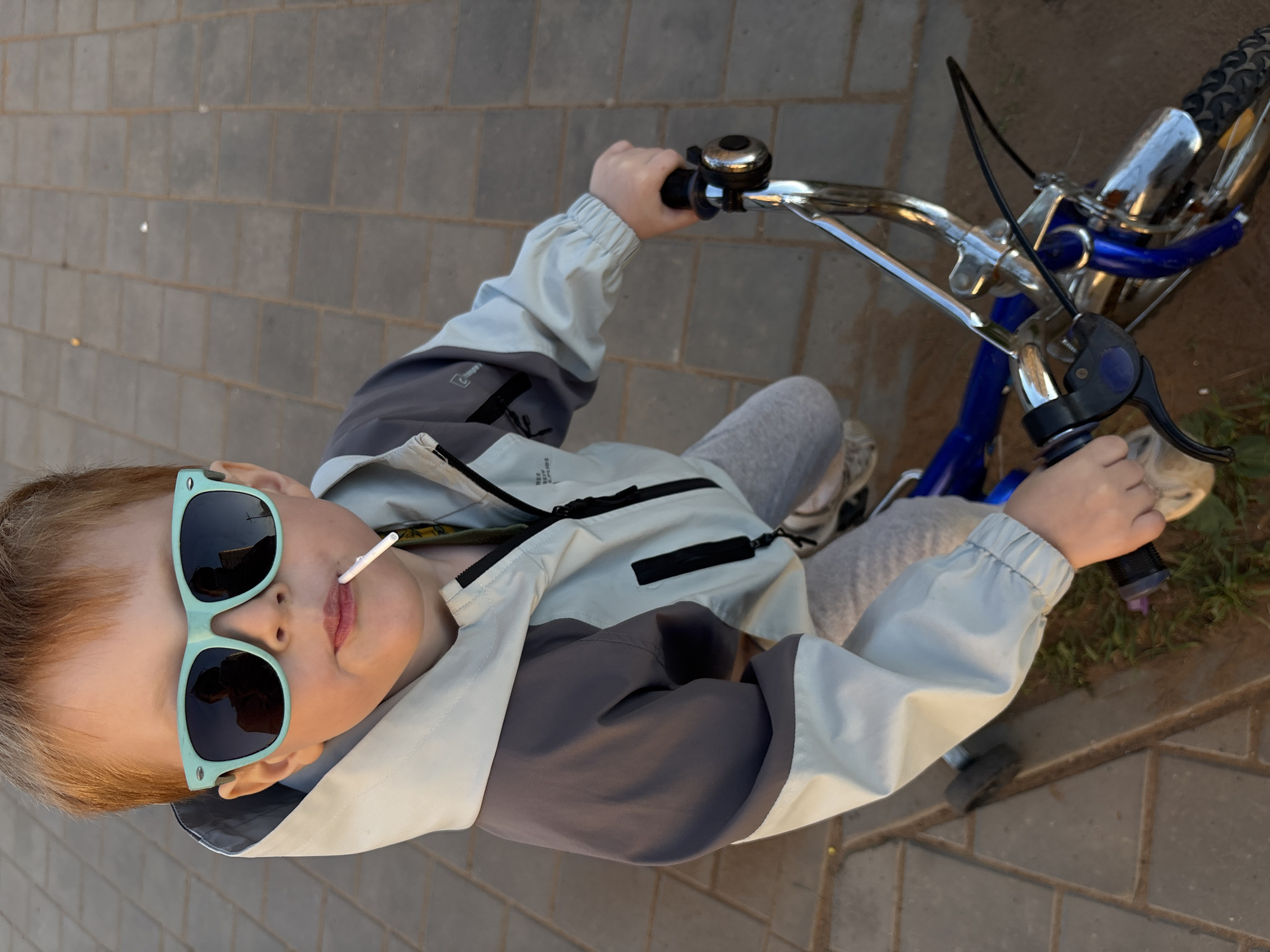 A young child in light gray sweatpants, a light blue jacket with dark gray accents, and teal sunglasses is riding a small blue bicycle. The child has a lollipop in their mouth. The photograph is taken from directly above, showing the child, the bicycle handlebars, and the front wheel, as well as the ground beneath. The ground appears to be a paved area with rectangular bricks. The location is not definitively ascertainable as Izhevsk, Russia from the given image.