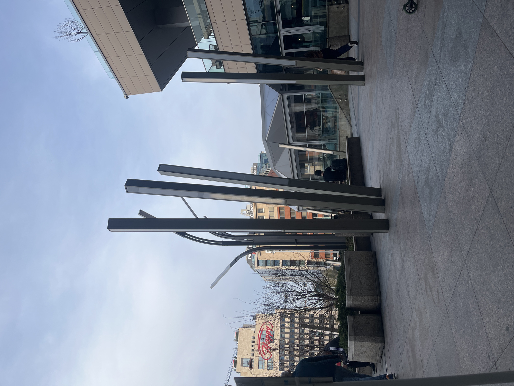 A ground-level, outdoor photograph shows a view of modern architecture juxtaposed with older buildings in Sofia, Bulgaria. Dominating the foreground are several vertical, grey rectangular posts with embedded lighting elements, likely part of a public art installation or architectural feature. These posts stand on concrete bases.
Behind these structures, buildings rise into the sky, including a large building bearing a prominent red “Happy” sign. The scene includes hints of other buildings with a mix of architectural styles, ranging from boxy modern structures with extensive glass facades to older buildings with more traditional detailing.
The sky is overcast and a bare tree is partially visible. People are present, but they are somewhat blurred and indistinct, suggesting they are in motion or the photograph was taken quickly. The ground is paved with large, grey tiles. The photograph captures a blend of the urban environment, with a focus on architectural elements and the presence of people within that setting.