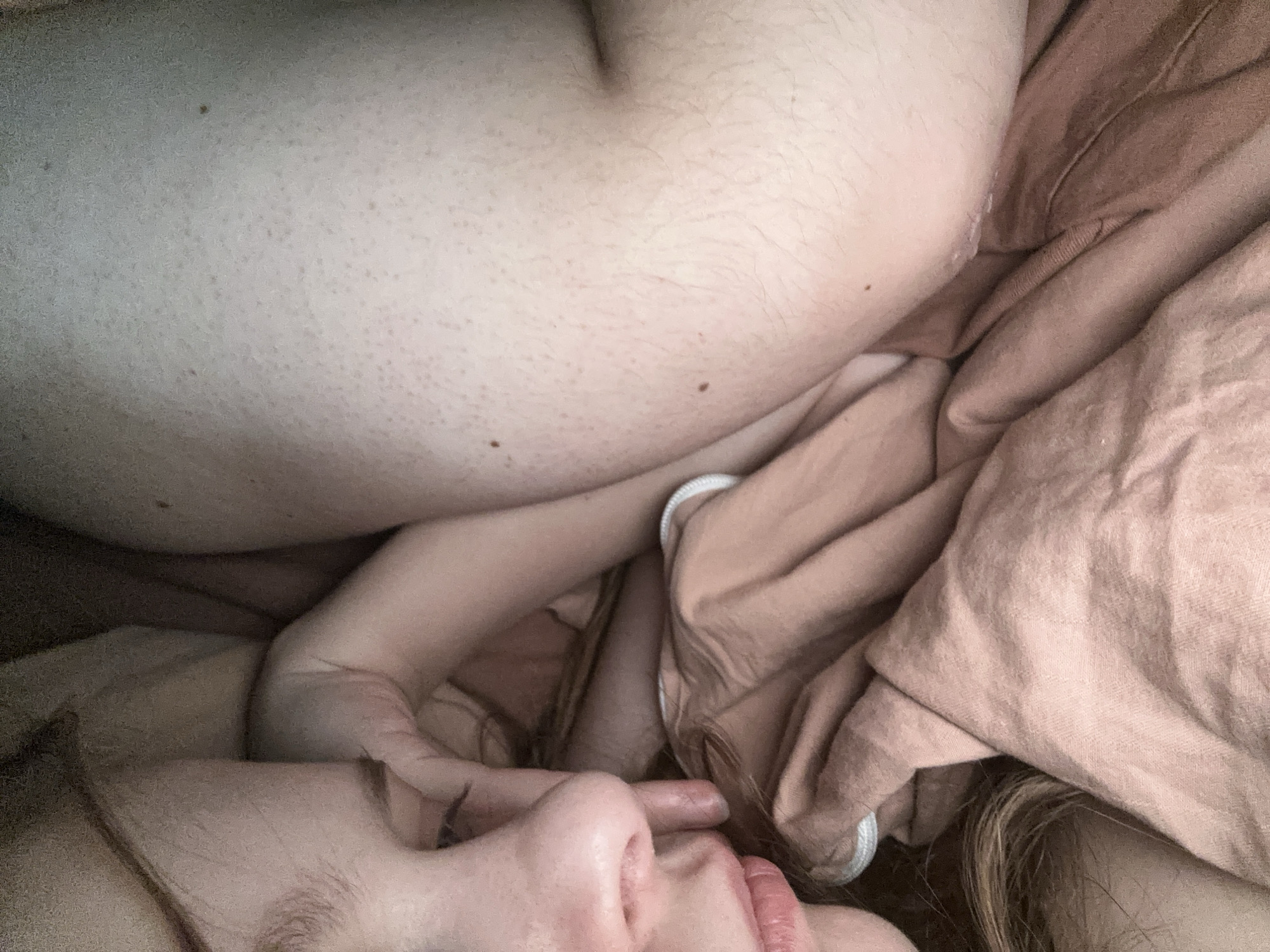 The image captures an intimate, close-up view of two individuals nestled among soft, crumpled fabric, likely bedding. The composition is tight, focusing on textures of skin, hair, and fabric.
In the bottom right quadrant, the profile of a person's face is visible. This includes a light-skinned nose, full lips with a light pink tint, and a chin. A portion of an eye is discernible, with long, delicate eyelashes. Strands of light brown hair frame the face. A hand or arm, also light-skinned with fine hair, is positioned near the face, possibly resting on it or interacting gently.
The upper left portion of the image reveals a significant area of bare, light-toned skin, appearing to be a torso or back. This skin exhibits fine, light body hair spread across its surface, along with several small, dark moles or freckles of varying sizes. The contours of the skin suggest a relaxed posture, with subtle dips and curves. A faint crease is visible towards the top center of this skin area.
Throughout the scene, a substantial amount of crumpled fabric in a muted reddish-brown or pinkish-tan color is present. This material appears soft and textured, consistent with bedsheets or a blanket, and is draped around and between the individuals, creating a sense of warmth and enclosure. A small white elastic band or cord is visible peeking out from beneath the fabric in the central area of the image.
The lighting is soft and diffused, casting gentle shadows and highlights that emphasize the tactile quality of the skin and fabric. The overall mood conveyed is one of intimacy, closeness, and quiet repose, suggesting a moment of rest or affectionate embrace. No text is visible within the image.