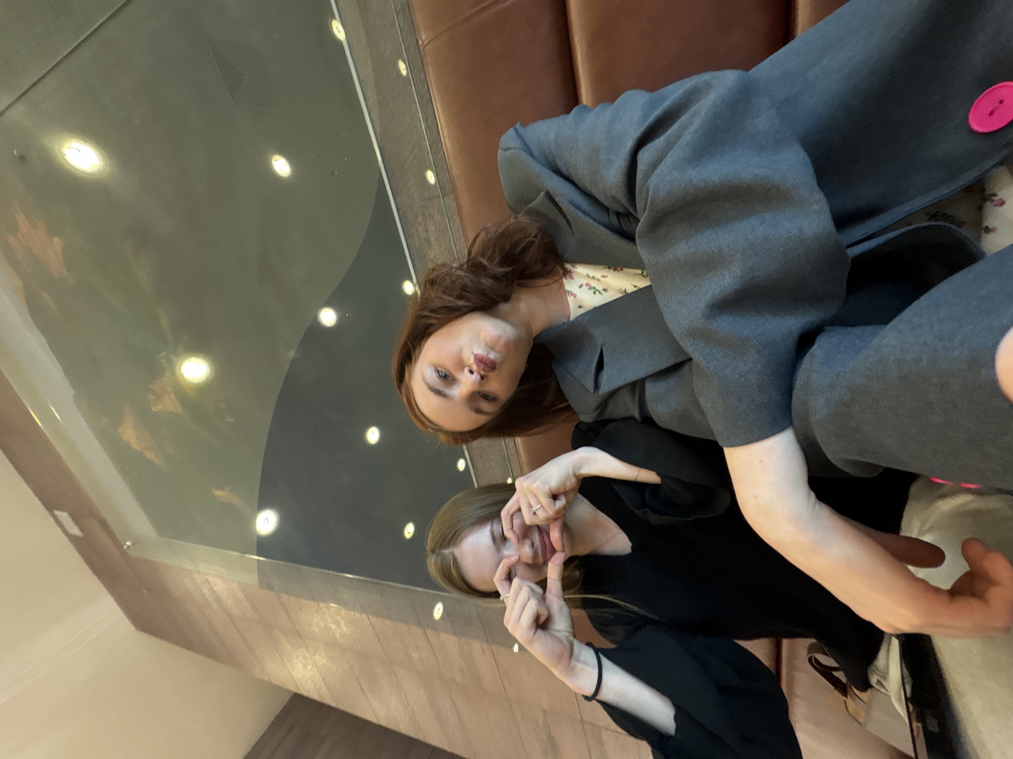 Two young women are captured indoors in a casual, playful pose. The image, originally rotated, shows them seated on a brown leather sofa.
The woman on the left (with reddish-brown hair) is looking directly at the camera with a pursed-lip expression. She wears a grey jacket, which features a bright pink button on the cuff, over a light-colored floral top.
The woman on the right (with blonde hair) is making a silly face, pulling at her cheeks with her hands and forming a heart shape with her fingers. She is wearing a black top and rings on her fingers, and a black elastic band is visible on her left wrist.
The background features a dark, reflective surface, likely a table or floor, showing reflections of several bright, circular overhead lights. Architectural details, including a wooden paneled wall and a glass partition, are visible, suggesting a modern indoor setting such as a cafe, lounge, or shopping center. The overall mood is lighthearted and candid. No visible text is present in the image.
