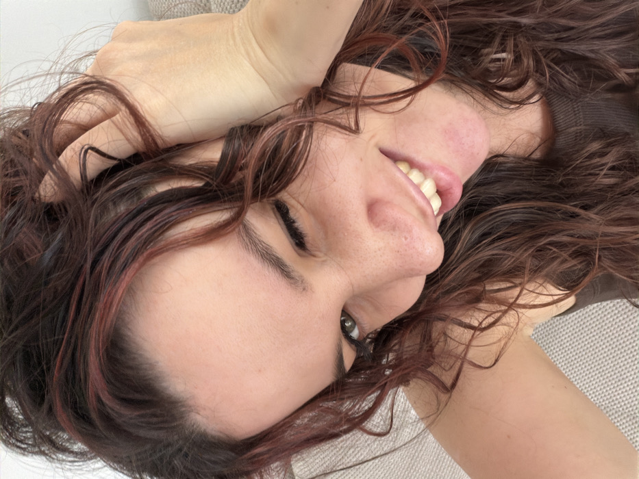 The image displays a close-up, high-angle portrait of a single individual, a female, with long, wavy dark brown hair featuring auburn highlights. Her right hand is visible in the upper right quadrant of the frame, with fingers intertwined in her hair. She is smiling broadly, exposing her teeth, while her right eye is squinted or winking, and her left eye is open. Her skin tone is fair, with visible freckles distributed across her nose and cheeks. Dark eyeliner is present on her upper left eyelid. She is wearing a dark brown or black ribbed top. The background is out of focus, showing a light-colored, textured fabric object, likely a piece of furniture, positioned to her left and below, against a plain light-colored surface or wall behind her head. No discernible elements within the image allow for the identification of a specific location in Budva, Montenegro.