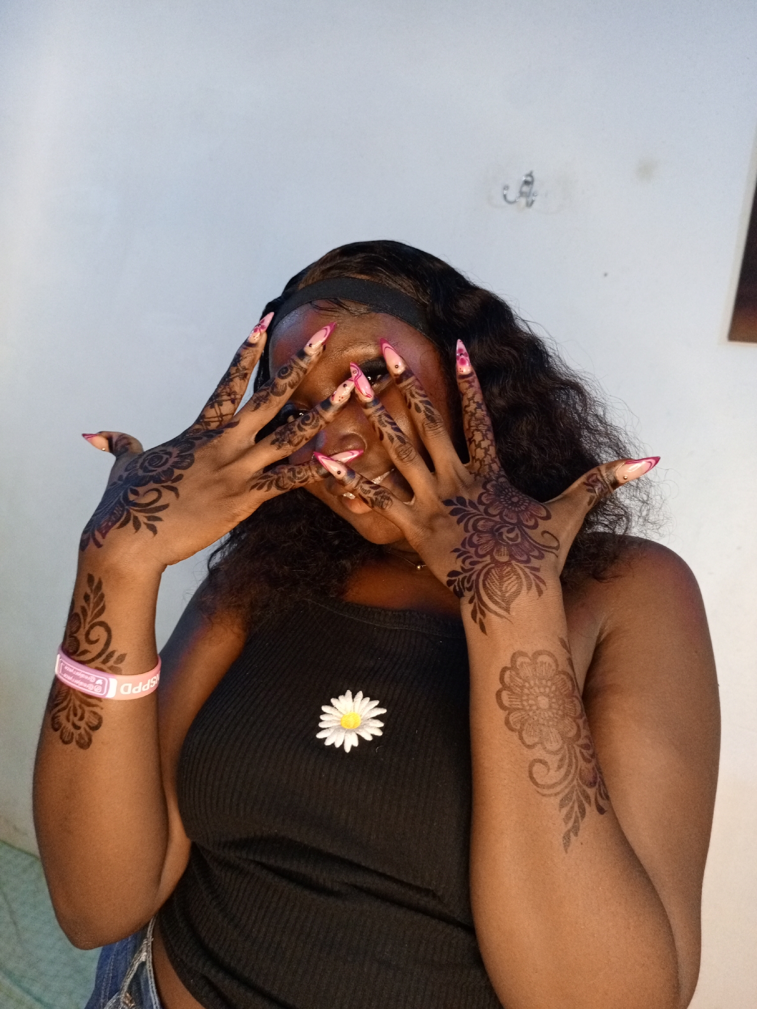 A person stands in front of a light-colored wall. The individual is wearing a black tank top with a white daisy attached to it. The person has long, dark curly hair, styled with a black headband. Intricate henna designs adorn both hands and forearms. The person's hands are raised, partially obscuring their face. The fingernails are long with a pink and white design. A pink wristband with lettering is visible on the person's left wrist.