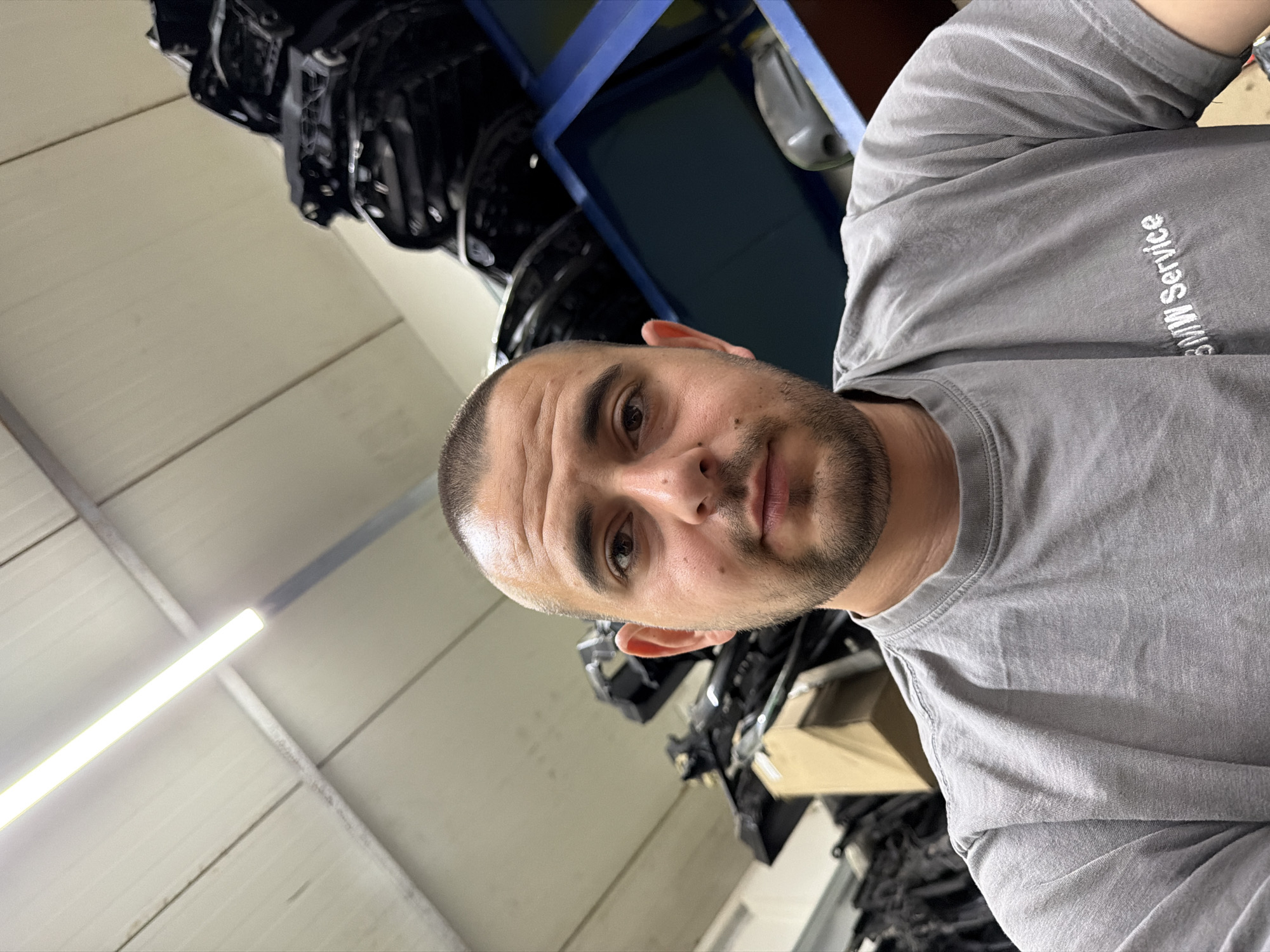 A young man with a buzz cut and a beard is taking a selfie in a warehouse. He is wearing a gray t-shirt with "BMW Service" embroidered on it. Behind him, shelves are stacked with car parts, including what appear to be car doors or body panels. The warehouse has a corrugated metal ceiling with fluorescent lighting. The location is a BMW service center in Burgas, Bulgaria.