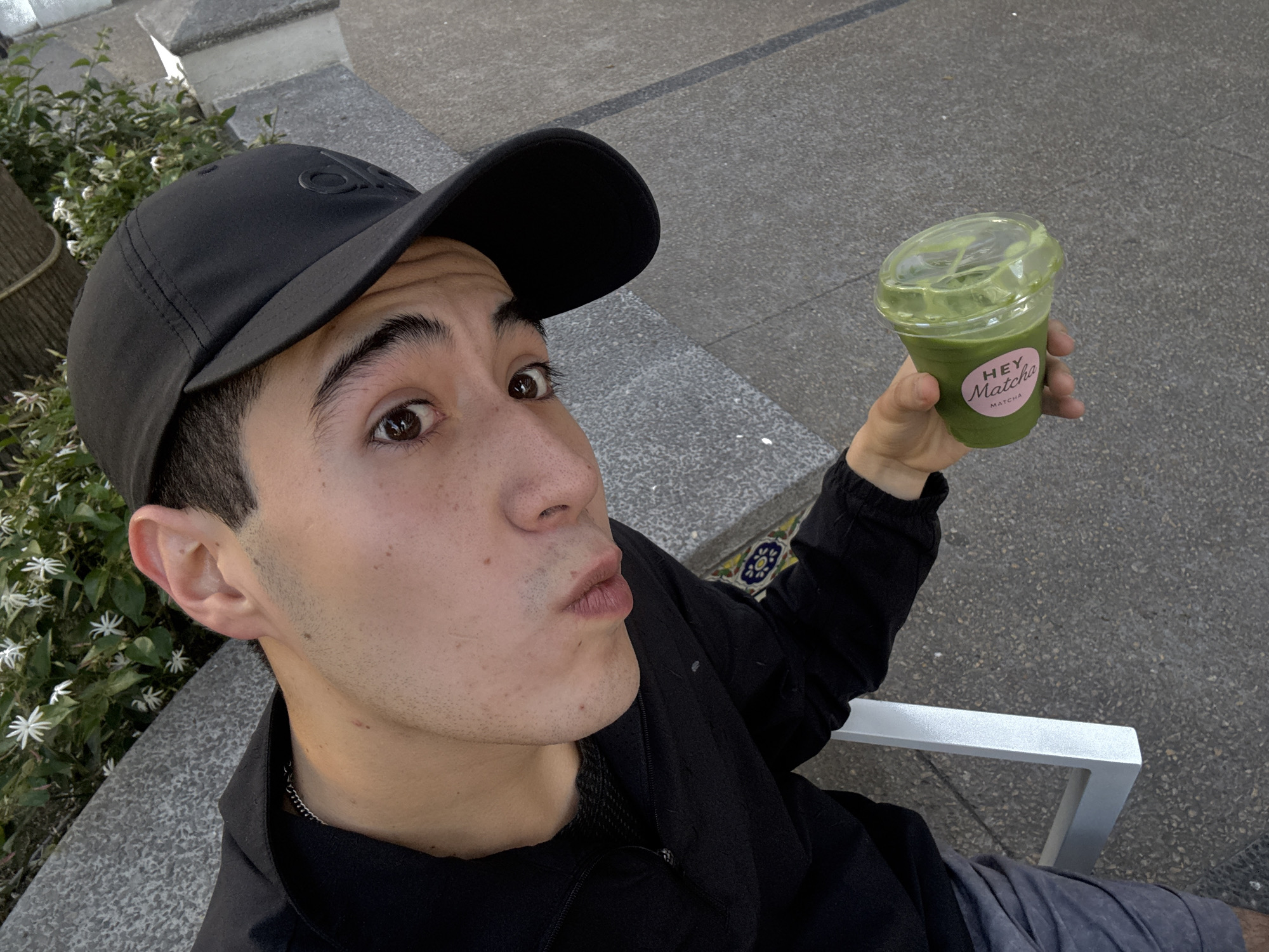 The image depicts a young man holding a cup of matcha in an outdoor setting. He is wearing a black baseball cap and a black jacket, and appears to be sitting on a bench. The cup has a "Hey Matcha" sticker on it. The background includes a stone surface and some foliage. The location, San Bernardino Tlaxcalancingo, Mexico, is not evident within the image itself, but is provided separately.