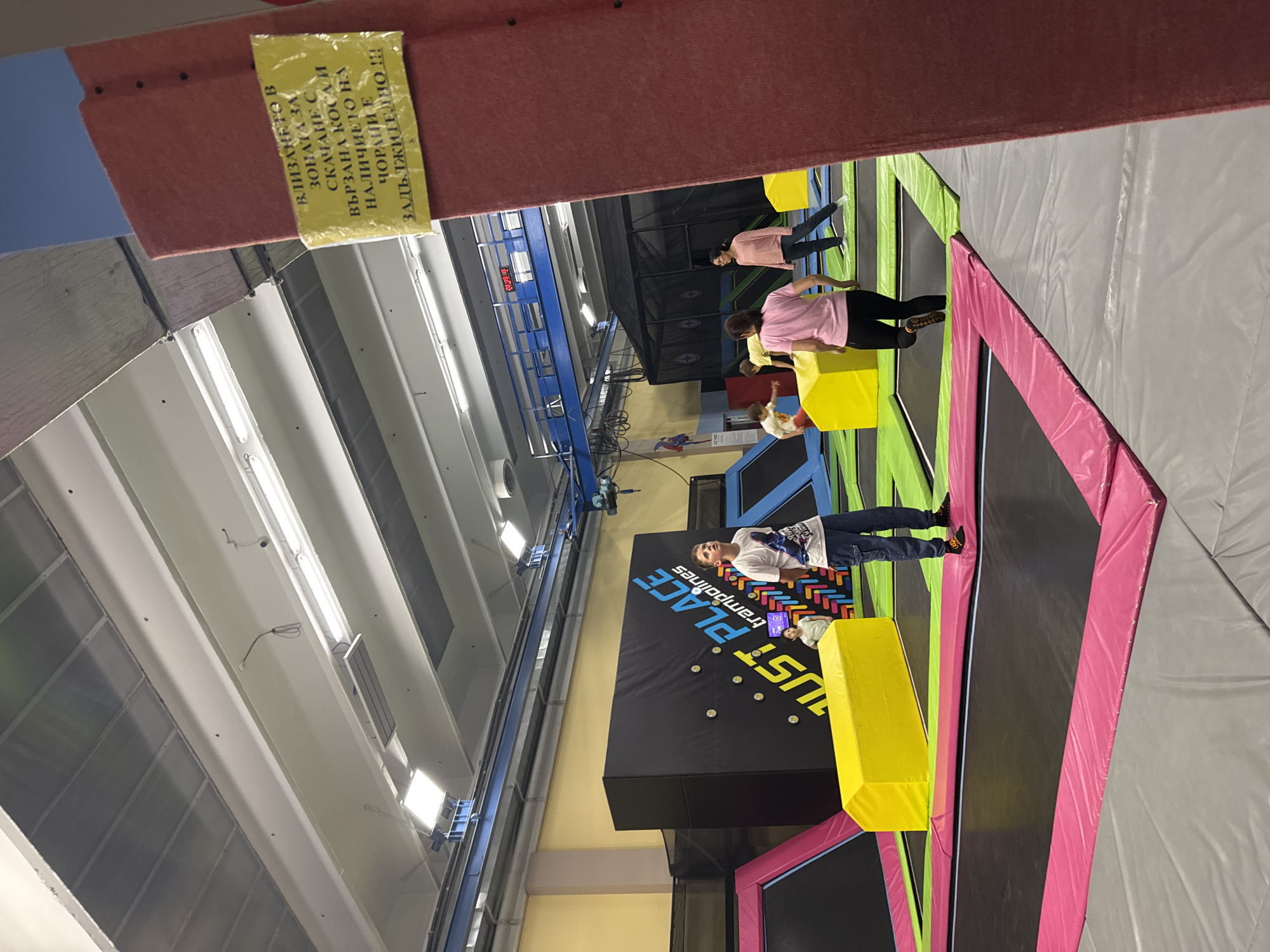 The image shows an indoor trampoline park, possibly "Just Place Trampolines", filled with trampolines and yellow foam blocks. Several people are present, some standing on the trampolines and one jumping on the trampoline. The building has a high ceiling with fluorescent lighting. A sign in Bulgarian script is visible, indicating that tied-up hair and socks are mandatory in the area. The location could be in Sofia, Bulgaria, based on the Bulgarian text on the sign.