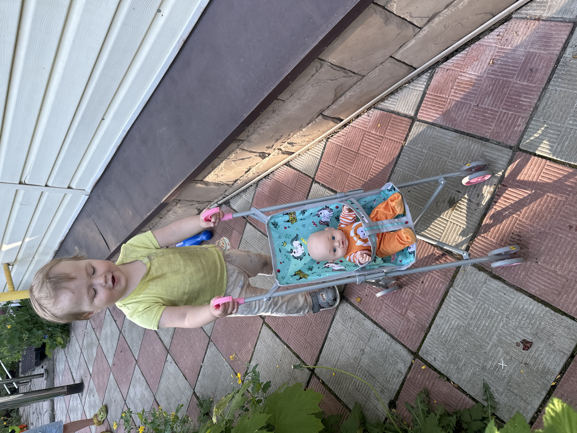 A young child with light hair pushes a toy stroller containing a doll on a paved surface. The child wears a light green shirt and beige pants. The stroller is silver with a blue patterned seat, and the doll is dressed in orange. The paving consists of alternating red and gray square tiles. A building with white siding and a dark brown trim is visible in the background. Green plants are visible at the bottom of the frame. The location is likely a residential area in Nizhny Novgorod, Russia, based on the appearance of the buildings and paving.