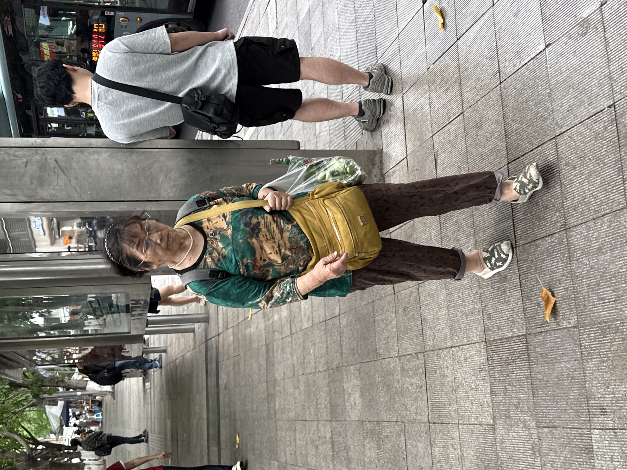 The image shows a street scene in what is likely Liangjiang, China, evidenced by the architecture and signage. An elderly woman with glasses and a headband walks along the sidewalk, carrying a plastic bag containing leafy produce and a yellow purse. She wears a patterned top with a green velvet-like jacket, brown pants, and patterned flat shoes. She is also wearing a pearl necklace. To her left, a man in a grey t-shirt, black shorts, and grey shoes, with a black messenger bag, stands near a bus. The environment includes a bus stop and a sidewalk made of grey tiles. Other pedestrians are visible in the background. A fallen leaf is seen on the sidewalk near the woman.