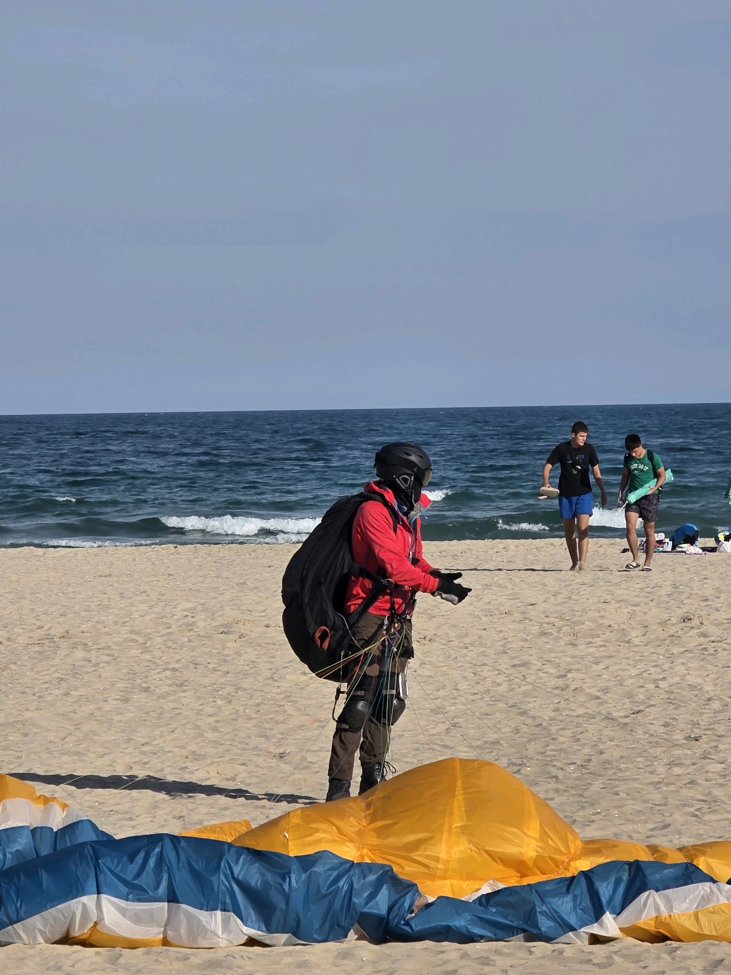 In Obrochishte, Bulgaria, on a sandy beach with the dark blue ocean in the background, a paraglider is preparing for flight. The paraglider, wearing a red jacket, black helmet, and dark pants, stands facing left with a large black backpack strapped to them and colorful paragliding equipment spread out on the sand in front. Two young men in casual attire walk along the beach further back, and other people and beach items are visible in the distance. The sky is a pale blue with a hint of clouds.