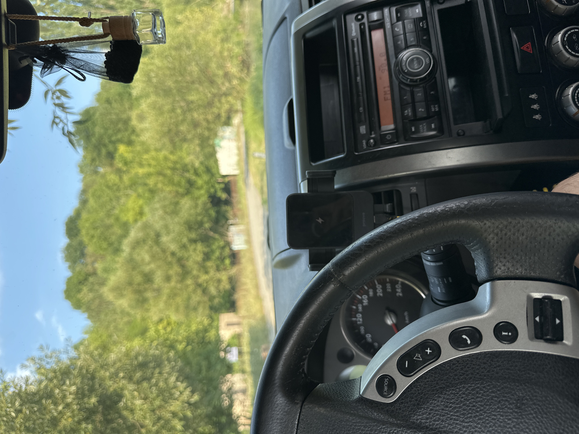 The image shows the interior of a car parked on a rural road, with a view of trees and a blue sky through the windshield. The dashboard includes a steering wheel with controls, a car radio, and a mobile phone holder. A car air freshener hangs from the rearview mirror. The scene is set in the village of Makiv, Ukraine.