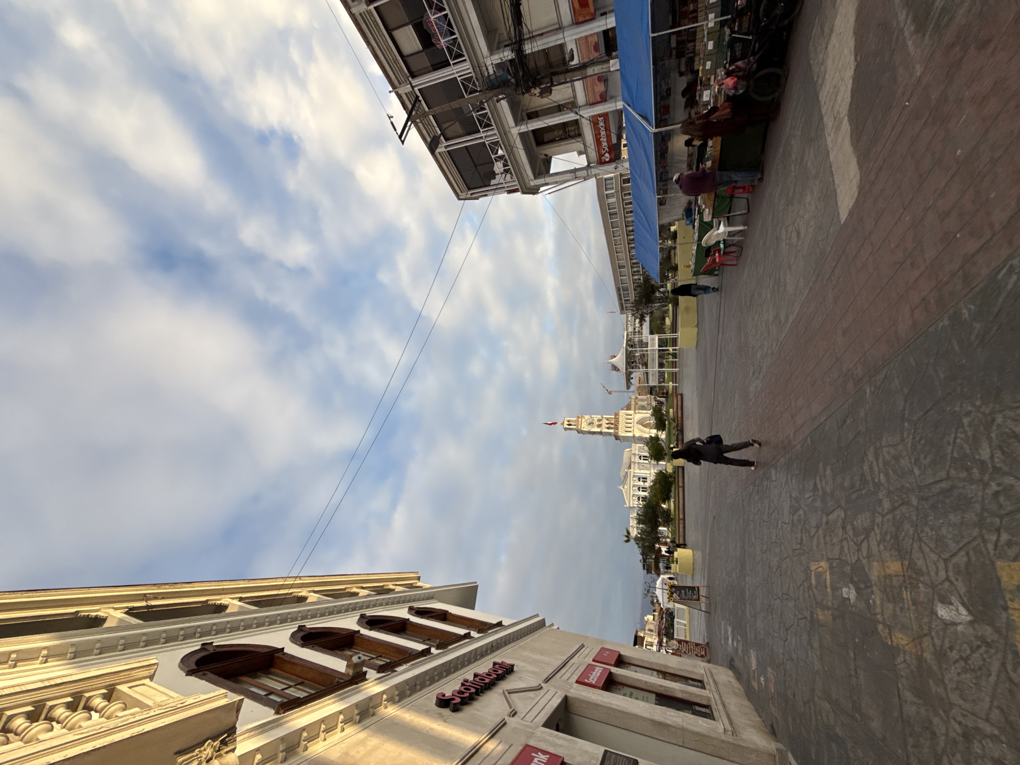 The image captures a view of the Plaza Arica in Iquique, Chile, during what appears to be the late afternoon or early evening. The foreground features a patterned cobblestone walkway leading into the plaza. To the left, a building with a light-colored facade is prominently featured, displaying signage for "Scotiabank" and "Banco Ripley." This building exhibits ornate architectural details, including carved elements and arched windows with dark wood frames.
The central and background elements of the scene reveal the heart of the plaza. A large, ornate building with a clock tower, likely the former Intendencia, stands at the far end. White, neoclassical buildings line the sides of the plaza, with trees and greenery interspersed. On the right side of the plaza, a row of market stalls with blue awnings is set up, displaying various goods. Several people are visible in the plaza. A woman in dark clothing is walking away from the camera towards the center of the plaza. Other individuals are browsing at the market stalls or walking in the distance. The sky above is partly cloudy with patches of blue visible, and the light suggests a low sun angle. Overhead electrical wires are strung across the scene.