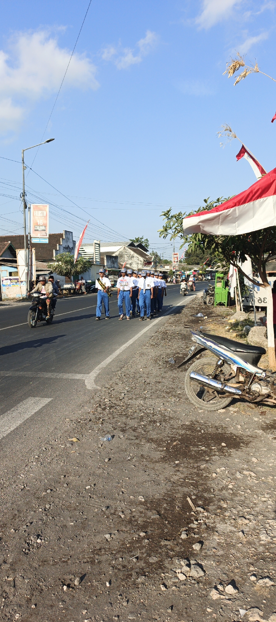 In the city of Kasiyan, Indonesia, a group of students in white shirts, blue pants, and caps are marching down a paved road under a clear blue sky. The students appear to be part of a school or youth organization, with some carrying sashes and others wearing numbered bibs. A motorcycle is visible in the background, ridden by two individuals, suggesting ongoing traffic on the road. The surrounding environment shows buildings with traditional Indonesian architecture, streetlights, and decorative red and white flags, possibly indicating a festive occasion or national celebration. The road itself is lined with gravel and dirt on the sides, with a discarded motorcycle on the right, adding to the streetscape. The scene captures a moment of community activity and daily life in an Indonesian town.
