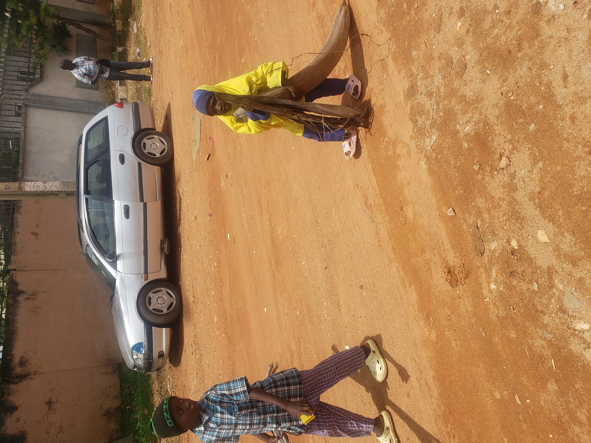 In Narayi, Nigeria, a young boy wearing a plaid shirt, checkered pants, and a green and black knit hat walks on a dirt road. He carries a yellow object in his right hand. To his right, another young child, dressed in a yellow headscarf and blue pants, holds a large piece of wood. In the background, a silver station wagon is parked, and a man in a checkered shirt and dark pants stands near a fence. The environment is dry and dusty with scattered debris on the ground.