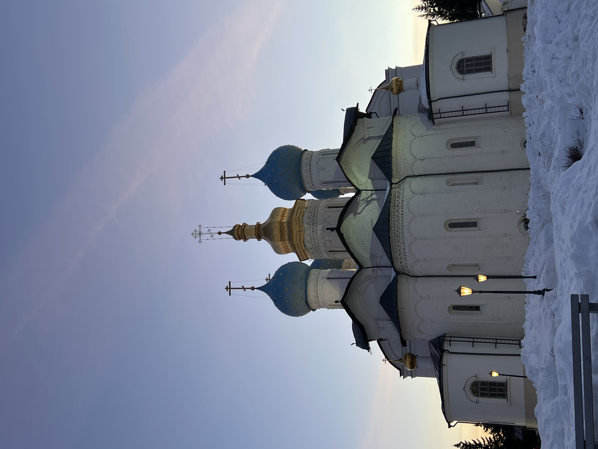 The image displays the Annunciation Cathedral, situated within the Kazan Kremlin in Kazan, Russia. The building is a multi-domed Russian Orthodox cathedral, predominantly white-walled with dark sloped roofs. A prominent central golden onion dome, topped with an Orthodox cross, stands between two smaller blue onion domes, each also bearing a cross. Additional smaller golden domes are visible on other sections of the cathedral. The structure features multiple arched windows. The ground in the foreground and around the cathedral's base is covered in deep, irregular drifts of snow, indicative of winter conditions. Three illuminated, traditional-style streetlamps are visible, standing among the snow in front of the cathedral. Coniferous trees are present in the background to the right. A dark-colored bench, partially obscured by snow, is visible in the lower left foreground. The sky exhibits a gradient from deep blue overhead to a lighter, yellowish-pink hue near the horizon, suggesting either dusk or dawn. No individuals are depicted.