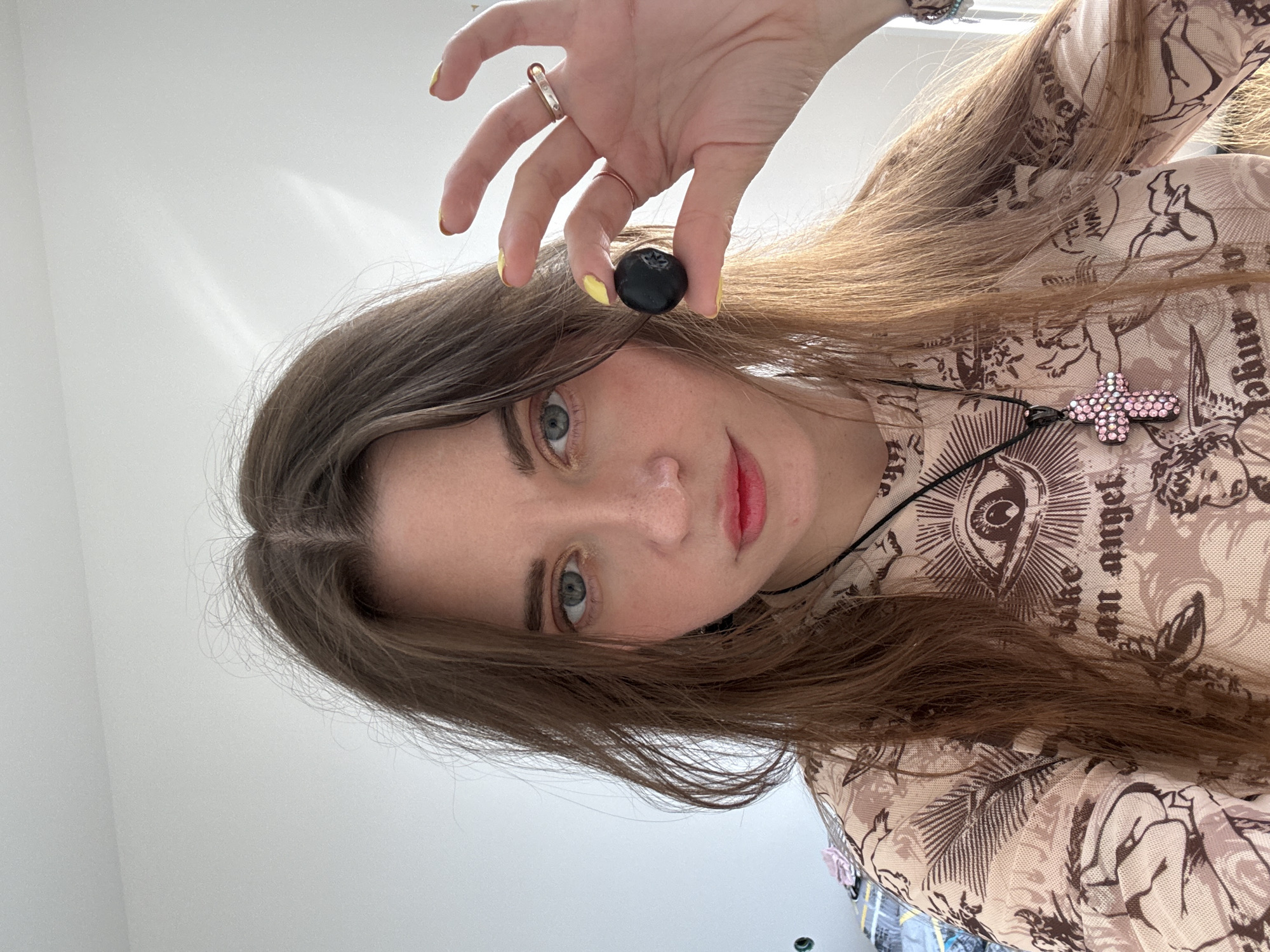 A woman with long brown hair holds a dark berry between her fingers. She is wearing a patterned shirt and a necklace with a pink cross pendant. The image is taken from a low angle, looking up at her. The background is a plain white wall. The location cannot be determined from the image.