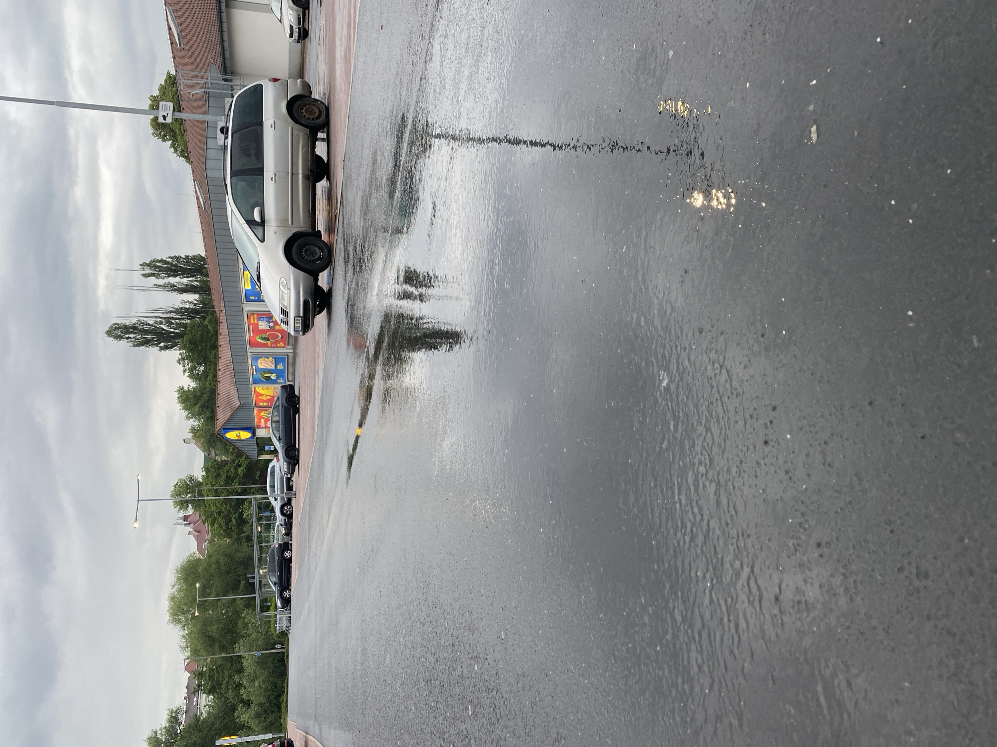 The image depicts a wet asphalt parking lot outside a Lidl supermarket in Bílina, Czechia. The sky is overcast and appears to have recently rained, as evidenced by the large puddles reflecting the surrounding environment. Several cars are parked in the lot, including a silver minivan in the foreground and a dark sedan further back. A shopping cart corral is visible near the entrance of the supermarket, which has the distinctive Lidl logo. Trees and buildings can be seen in the background, suggesting a suburban or urban setting. The overall impression is one of a typical shopping day in a European town under cloudy weather conditions.