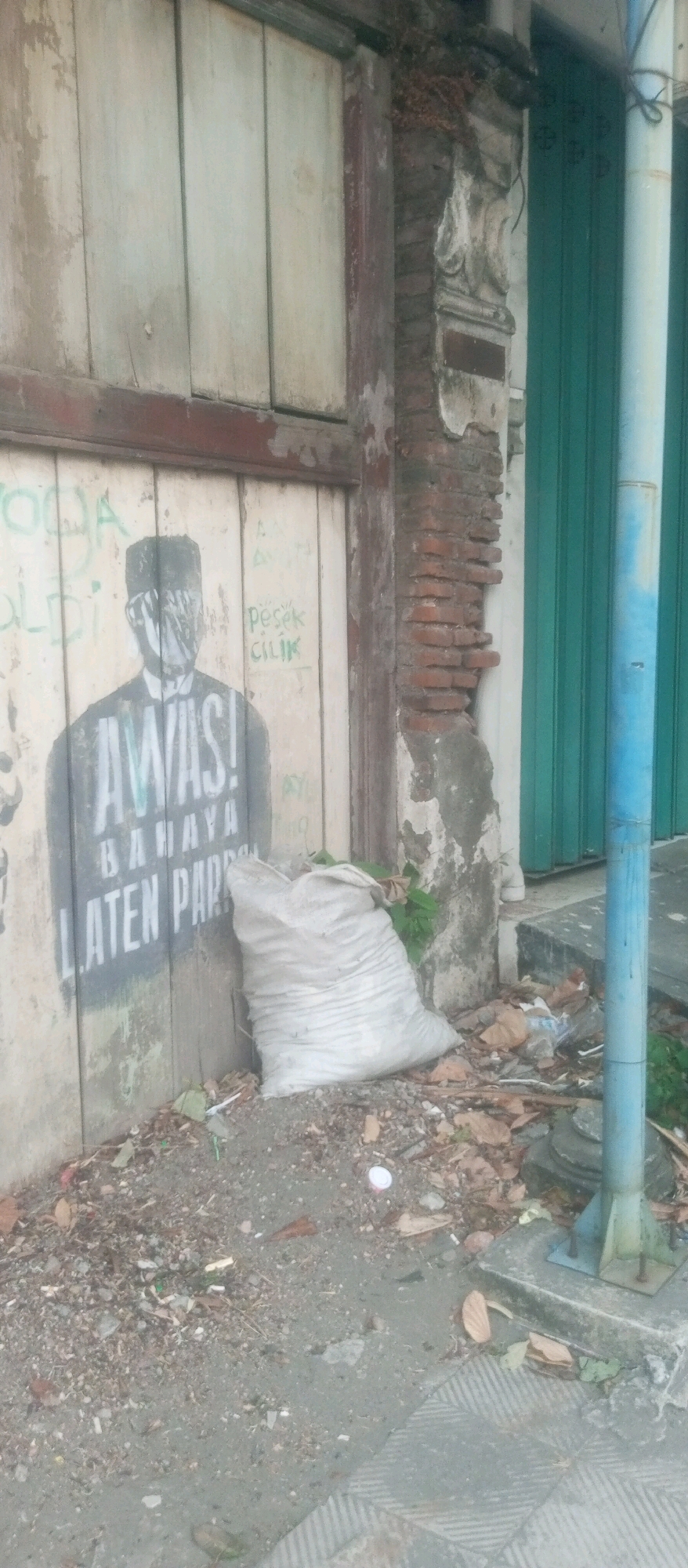The image captures a street scene in Kediri City, Indonesia, focusing on a weathered wooden door that has been spray-painted with graffiti. A stenciled image of a person wearing a fez and a shirt is prominent, with the word "AWAS!" ("Beware!") spray-painted in large, bold letters across the chest area. Below this, the words "BAHAYA" ("Danger") and "LATEN PAR?" are partially visible. To the right of the graffiti, a large, white, woven sack is leaning against the door, filled with what appears to be garbage or discarded materials. The ground in front of the door is littered with debris, including leaves, plastic wrappers, and cigarette butts. A blue metal pole stands to the right of the sack, next to a green metal shuttered door. The overall impression is one of urban decay and neglect.