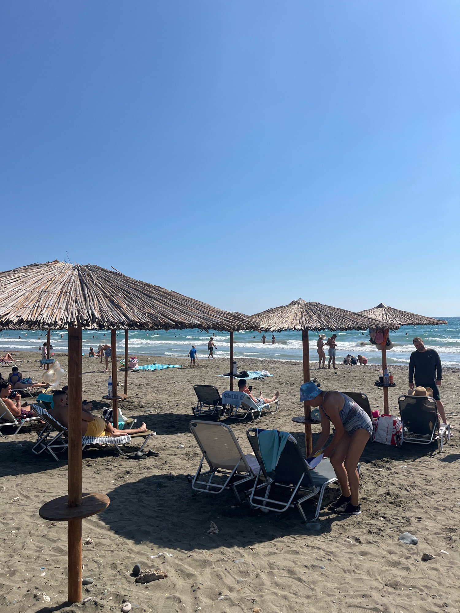 The image depicts a sandy beach with several thatched umbrellas providing shade to lounge chairs. In the foreground, a person wearing a patterned swimsuit and a blue bucket hat is tending to beach items near a lounge chair. To the right, a man in a dark long-sleeved shirt stands by another lounge chair. Further back, various individuals are scattered across the beach and in the shallow water, some seated on chairs, others wading or playing. The sea is visible with gentle waves under a clear blue sky. The location is identified as Episkopi beach in Episkopi Lemesou, Cyprus, not the United Kingdom.