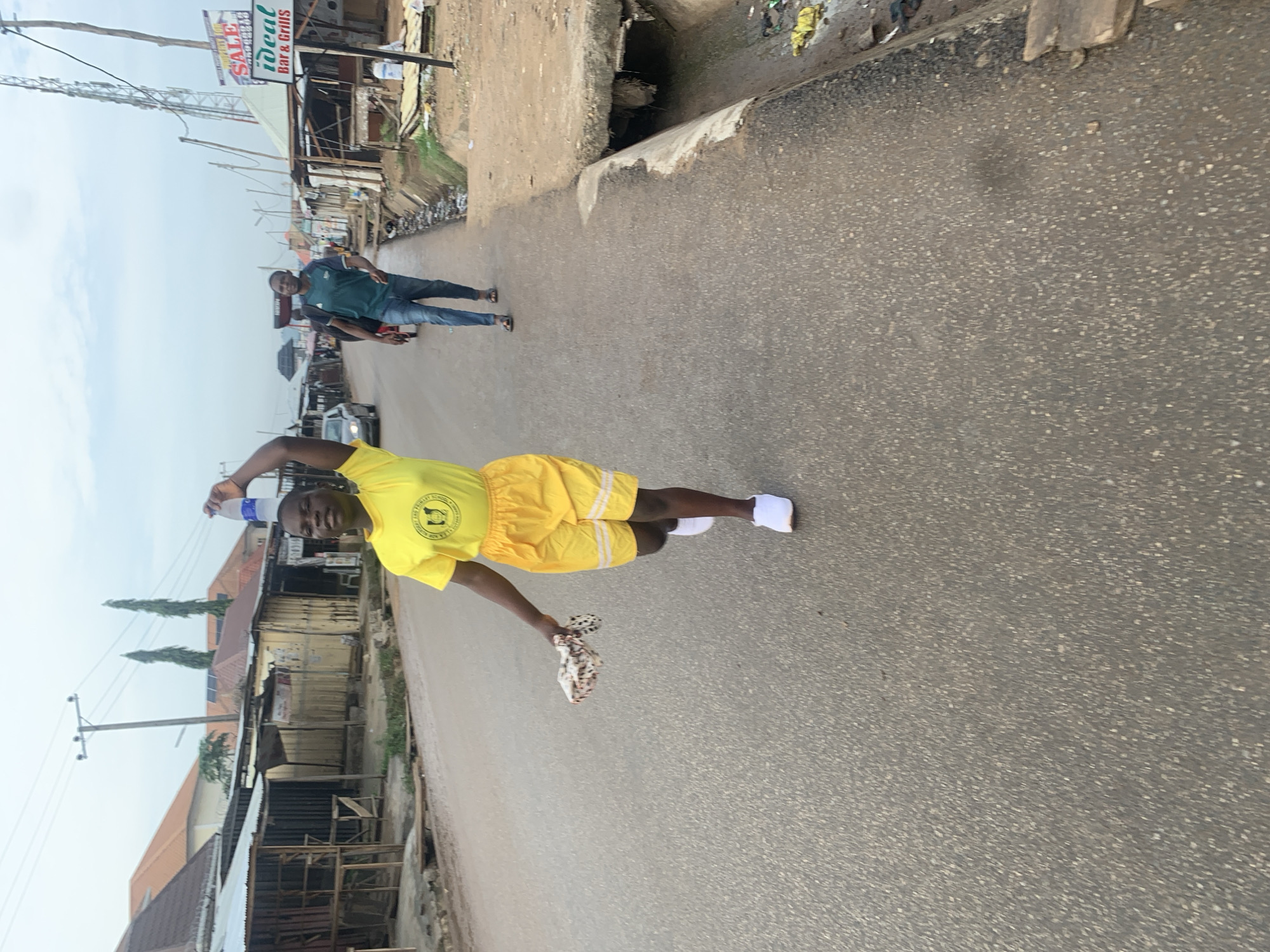 A young woman in a yellow t-shirt and yellow shorts is walking on a paved road in Abuja, Nigeria, with a plastic bottle balanced on her head. She is holding a patterned cloth in her right hand. Behind her, a man in a green t-shirt and blue jeans is also walking on the road. The street is lined with small businesses and residential buildings. The sky is overcast. The road appears to be in a commercial area, possibly a market district.