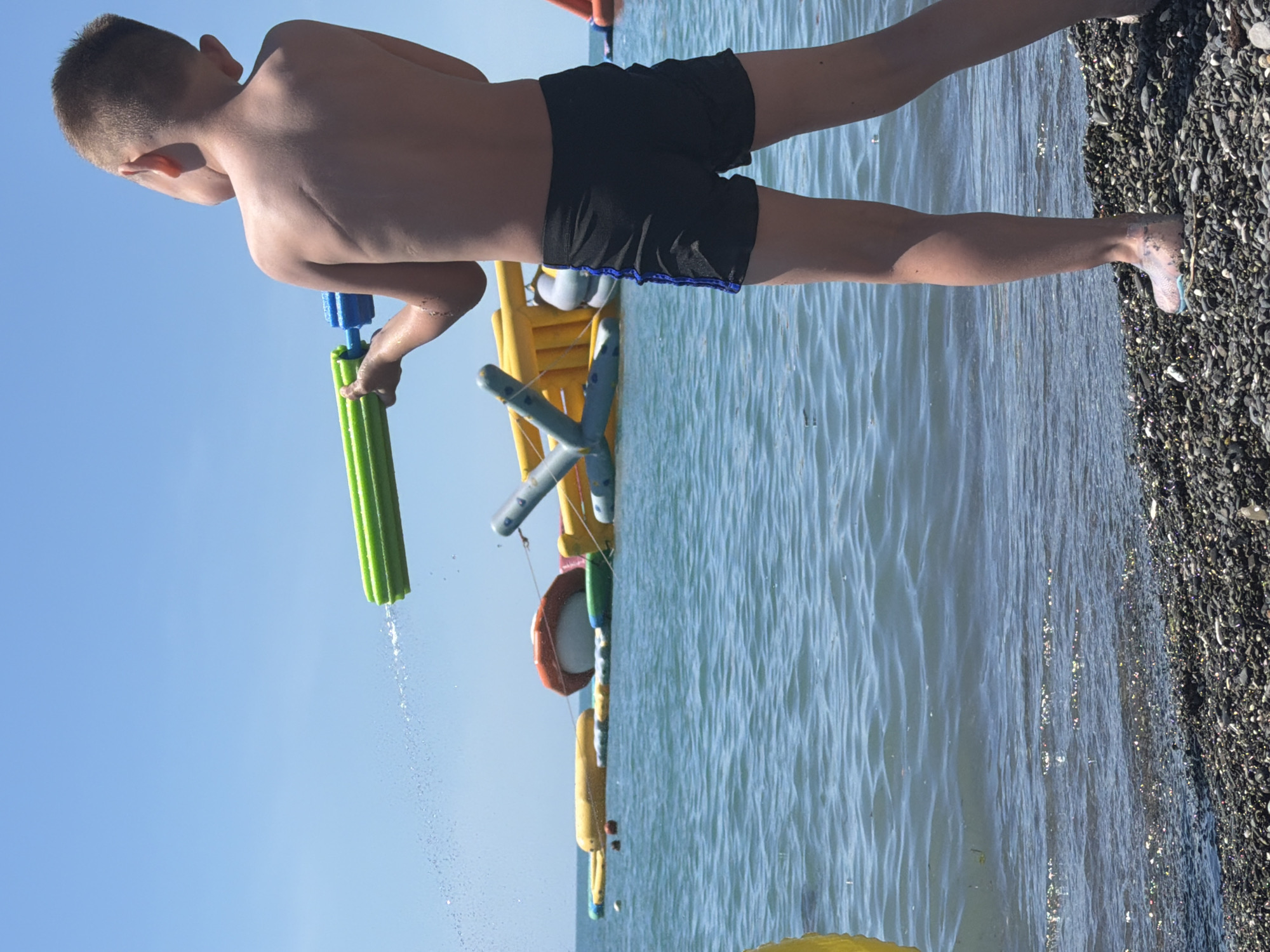 The image shows a young boy in black swim trunks holding a green and blue water gun, shooting a stream of water from it. He is standing on a pebble beach with the sea in front of him. In the background, there are several large inflatable structures in the water, which appear to be part of a water park. The sky is clear and blue. The location is identified as the city of Sochi, Russia.
