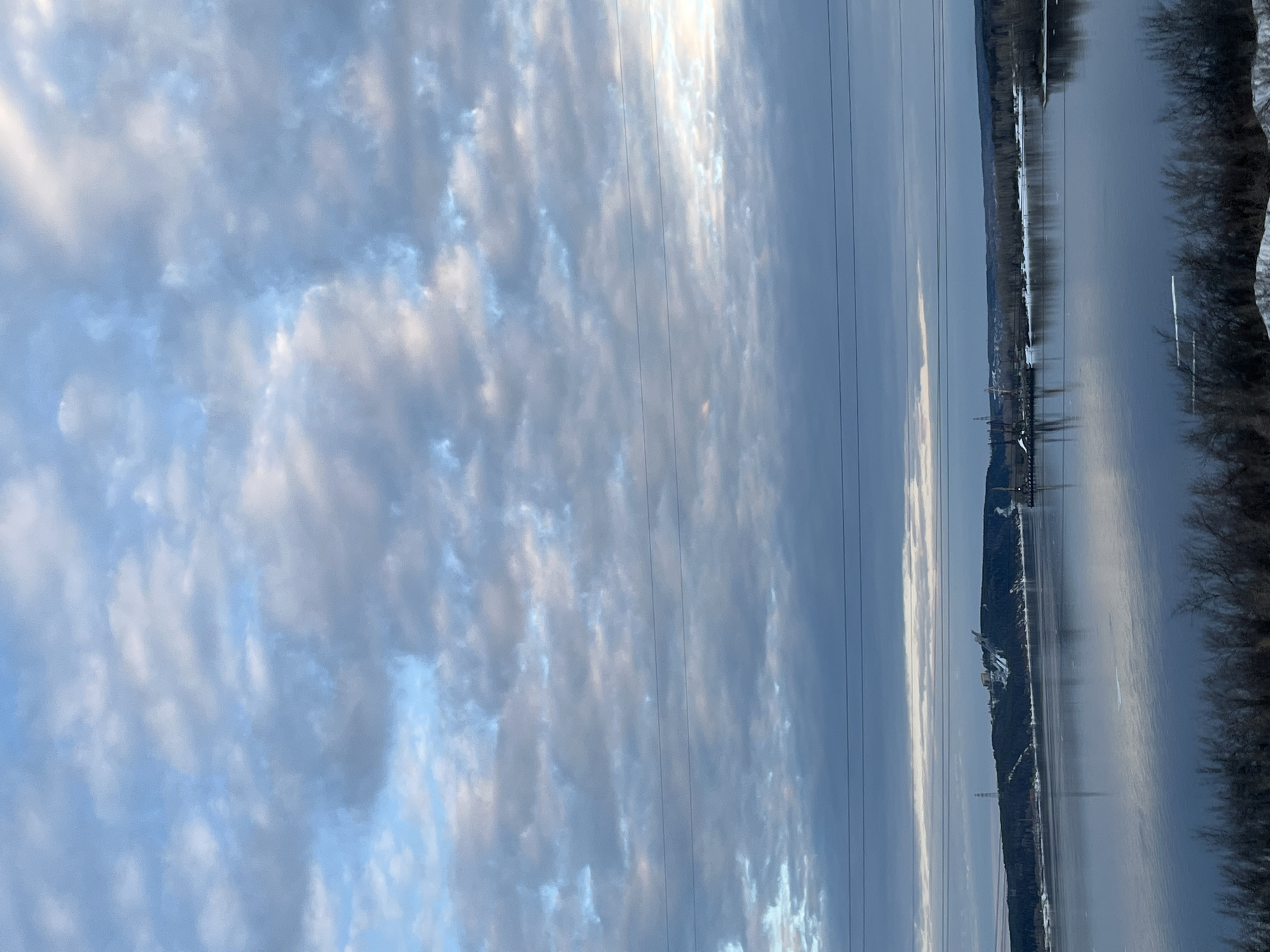The image depicts a panoramic view of Chaikovsky, Russia, featuring a river and an industrial complex. The sky is filled with textured clouds. Power lines run vertically across the frame. On the right, a body of water, likely the Kama River, reflects the sky. Along the riverbank, vegetation and snowy patches are visible. In the distance, the silhouette of an industrial area with structures and towers is discernible.