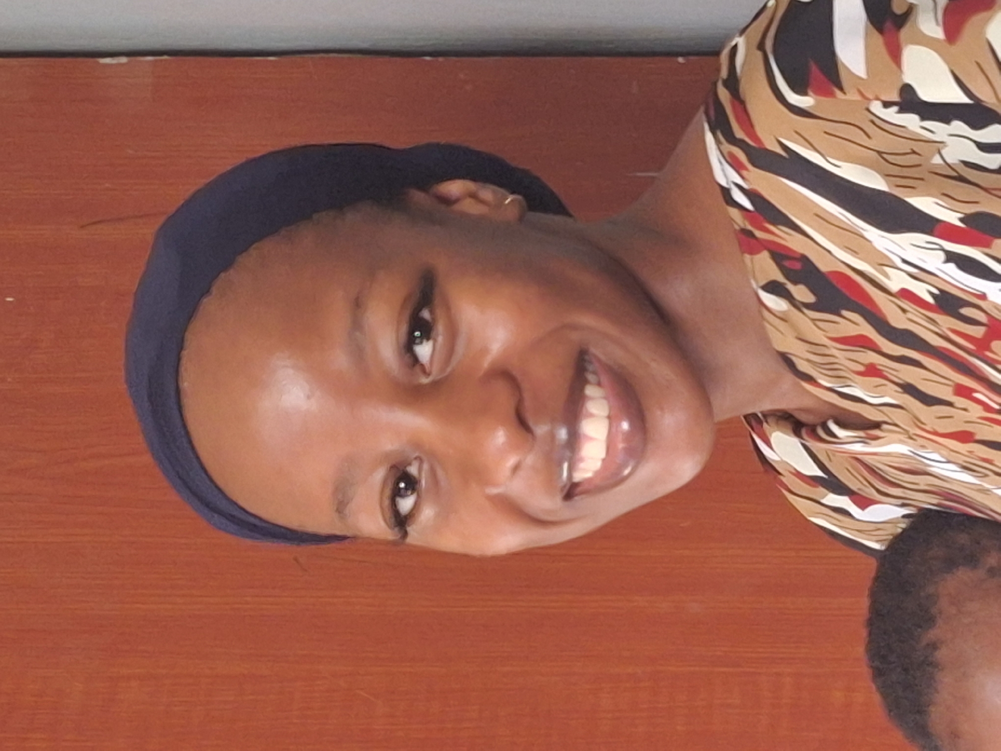 A portrait of a smiling dark-skinned woman wearing a navy blue head covering and a patterned tan, black, white, and red top. The background appears to be a reddish-brown wall. It is not possible to determine the exact location in Yelwa, Nigeria, or any details about subjects, objects, actions, or interactions from this image alone.