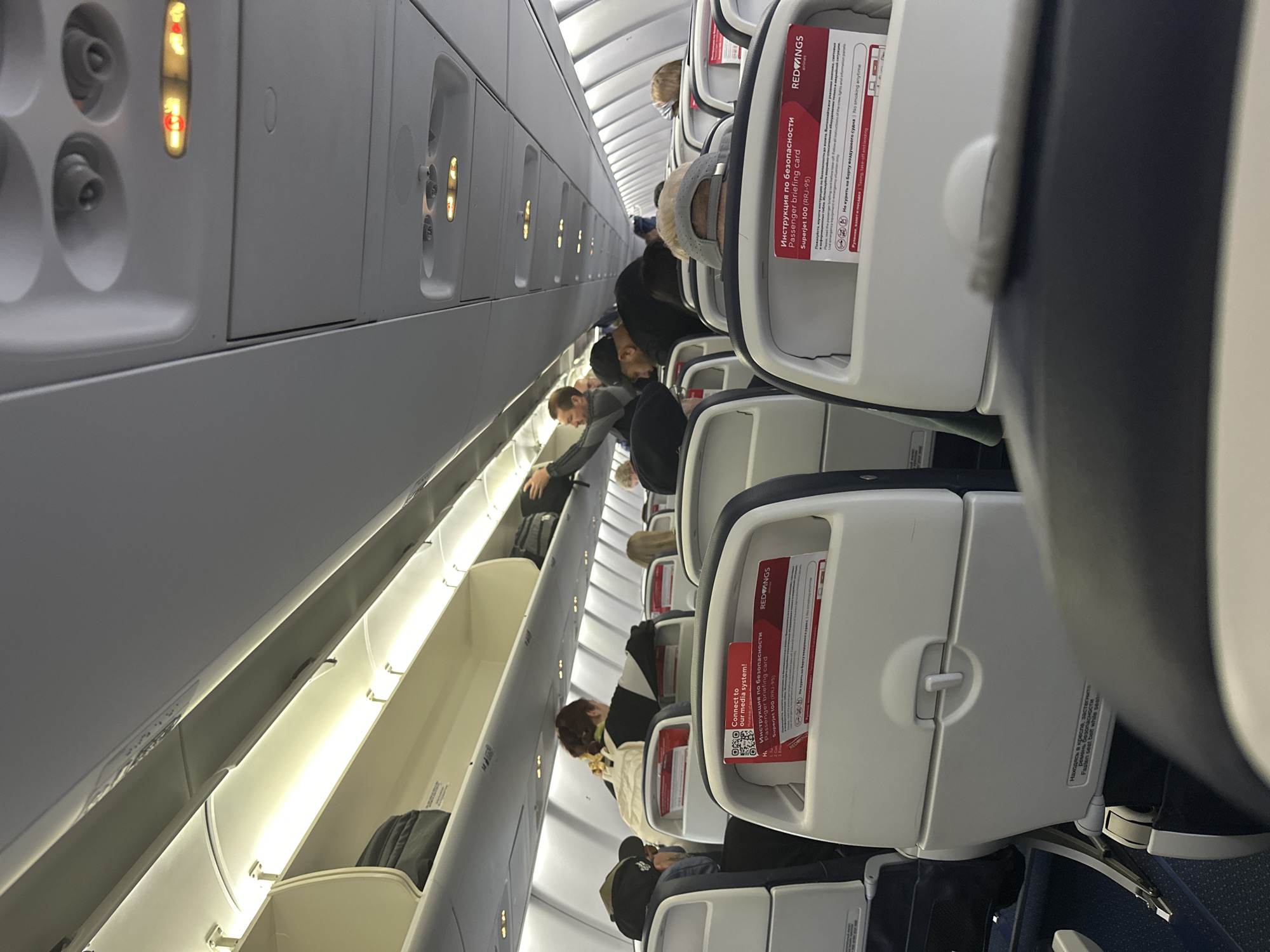 The image shows the interior of an airplane cabin. Passengers are seated in rows. Overhead compartments are visible above the seats. Each seat has a safety briefing card in the seat pocket. The card indicates the airline is "Red Wings" and the aircraft is a Superjet 100 (RRJ-95). The text on the card is partially in Russian. The lighting suggests it is daytime. The aircraft is of Russian origin. The location of the city of Sochi, Russia, is not visible.
