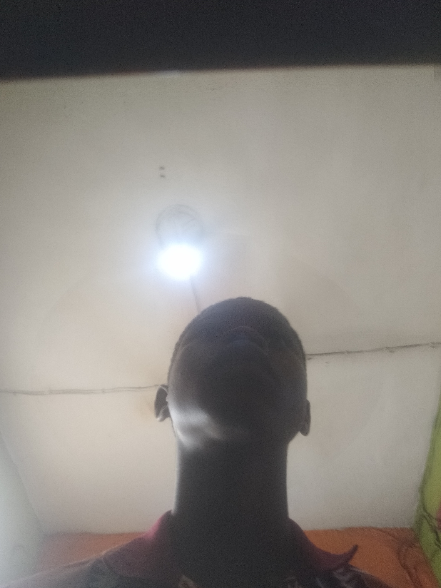 A low-angle photograph depicts a dark-skinned individual from the chest up, looking toward a bright ceiling light. The room appears to be dimly lit save for the light source, obscuring details of the person's face. The background suggests a room with off-white walls and indistinct items near the edges of the frame. Given the specified location of Shangisha, Nigeria, the setting is likely a residential interior.