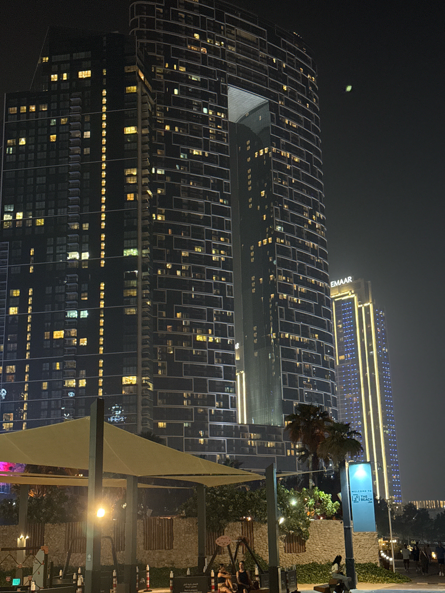 This image captures a night view of a waterfront area in Dubai, United Arab Emirates. The foreground features a beige awning supported by dark poles, providing shade over a seating area. Beyond the awning, a stone wall is visible, with some greenery and a swing set. A blue sign with white text reads "WELCOME TO THE BEACH JBR". Several people are present in the mid-ground and background, some seated on benches and others walking.
Dominating the background are two prominent skyscrapers. The taller building on the left is dark with numerous illuminated windows and features a distinctive angular cutout near its top. To the right of this building is a slightly shorter skyscraper with a lighter facade, illuminated by vertical lines of blue and yellow lights. The words "EMAAR" are visible in white lettering on this building. The sky above is dark, indicating it is nighttime, and a faint green light is visible in the upper right quadrant of the sky. The overall impression is of a vibrant urban night scene, likely a popular leisure destination within Dubai.