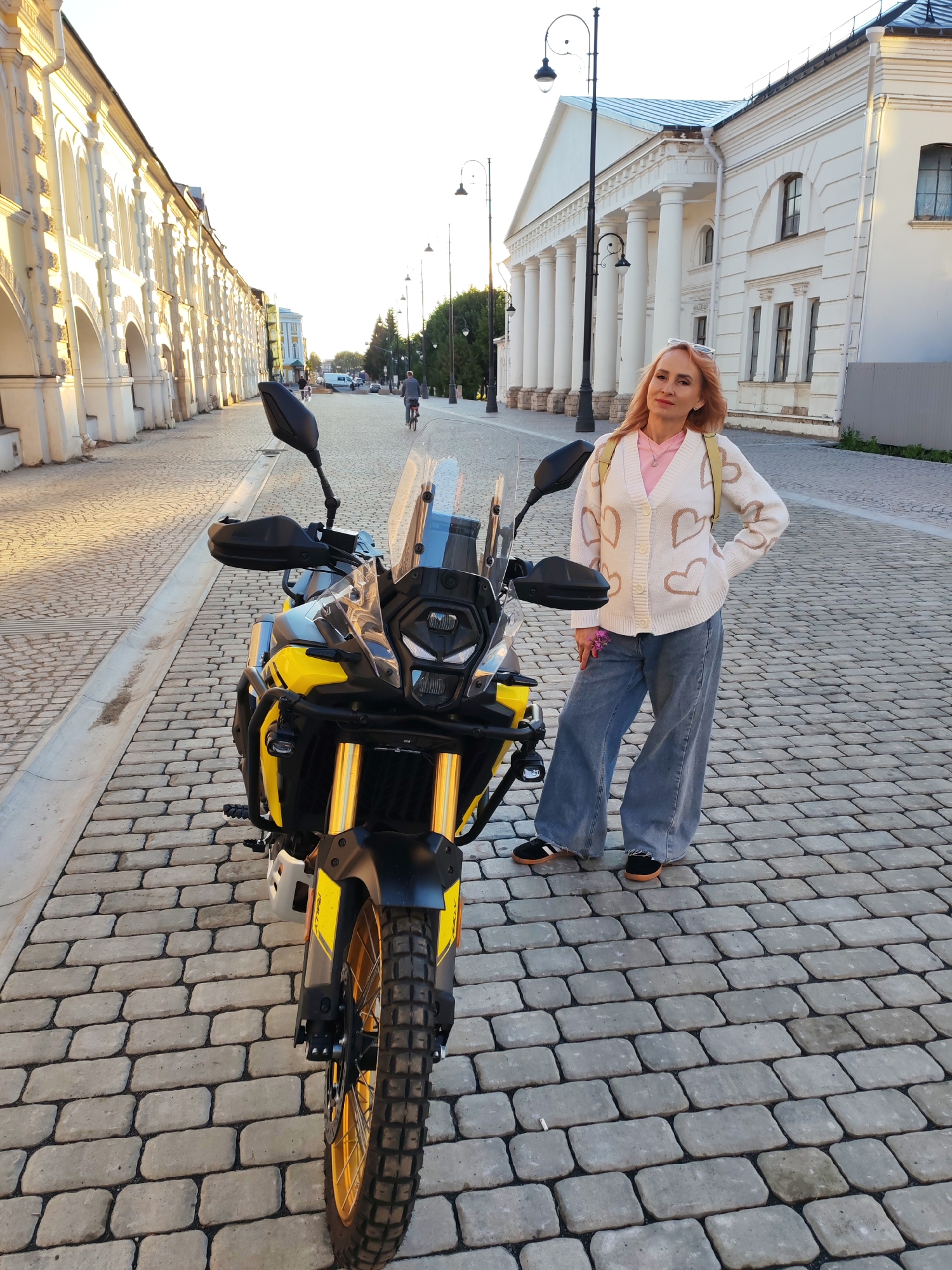 A woman stands on a cobblestone street in front of a yellow and black motorcycle. She is wearing a light-colored cardigan with heart patterns, a pink shirt, and wide-leg blue jeans. She has blonde hair and is wearing sunglasses on her head. The motorcycle is parked on the left side of the frame, facing slightly towards the viewer. In the background, there are classical-style buildings with columns and arched windows, typical of historic city architecture. The street is paved with light-colored cobblestones. The sky is bright and clear, indicating daytime, likely late afternoon due to the soft lighting. The overall setting appears to be a city square or historic street in Bol'shie Mkhi, Russia, given the architectural style and paved street.