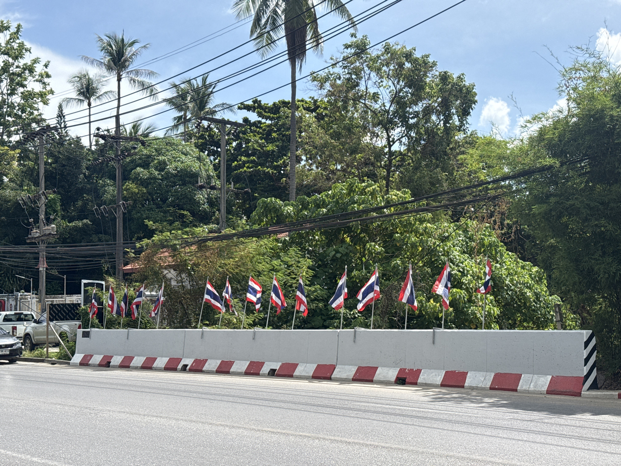 The image shows a street scene in Baan Chaweng Noi, Thailand. A line of Thai flags on poles is mounted on top of a concrete barrier. The barrier has red and white stripes at its base. In the background, lush green trees and palm trees are visible, with power lines strung across the scene. A white pickup truck and a darker colored car are parked to the left of the barrier, with a building partially visible behind them. The road in the foreground is paved. The sky is blue with scattered white clouds.