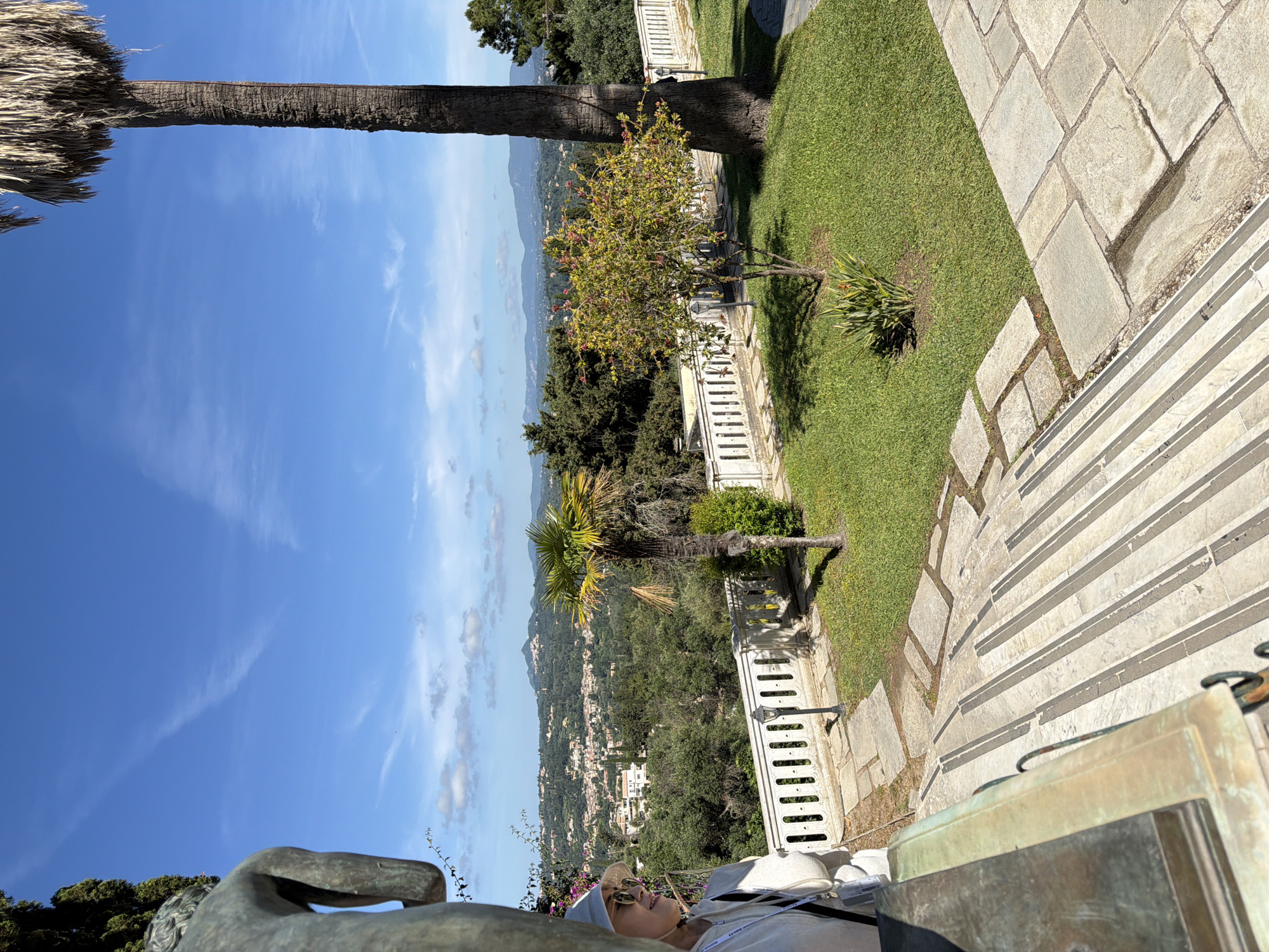 This image captures a scenic outdoor view from an elevated garden or terrace in Gastouri, Greece, under a clear, bright blue sky with a few scattered clouds. The dominant feature of the landscape is a distant valley or coastal area, characterized by rolling hills covered in lush green vegetation and a town or village nestled among them, identifiable by numerous white-roofed buildings.
In the foreground, a manicured garden is visible, featuring vibrant green grass and various trees. A tall palm tree with a textured trunk stands prominently on the left side, with its fronds partially visible. Another smaller tree, bearing pinkish-red blossoms, is situated in the mid-ground. Architectural elements include a light-colored stone pathway composed of irregularly shaped flagstones, and a white stone balustrade with a decorative streetlamp. Towards the lower left, part of a bronze-colored statue, depicting a human form, can be seen.
On the right side of the image, a person is shown relaxing, reclining in a chair or on a bench. They are wearing a light-colored hat and sunglasses, seemingly enjoying the expansive view. In the immediate foreground on the right, there's a large, square, bronze-toned object, possibly a planter or a statue base, next to a set of light-colored stone stairs with horizontal stripes.
The bright sunlight casts distinct shadows, suggesting it is midday or early afternoon. The overall scene conveys a peaceful and leisurely atmosphere, highlighting the natural beauty and well-maintained grounds. No discernible text is visible in the image.