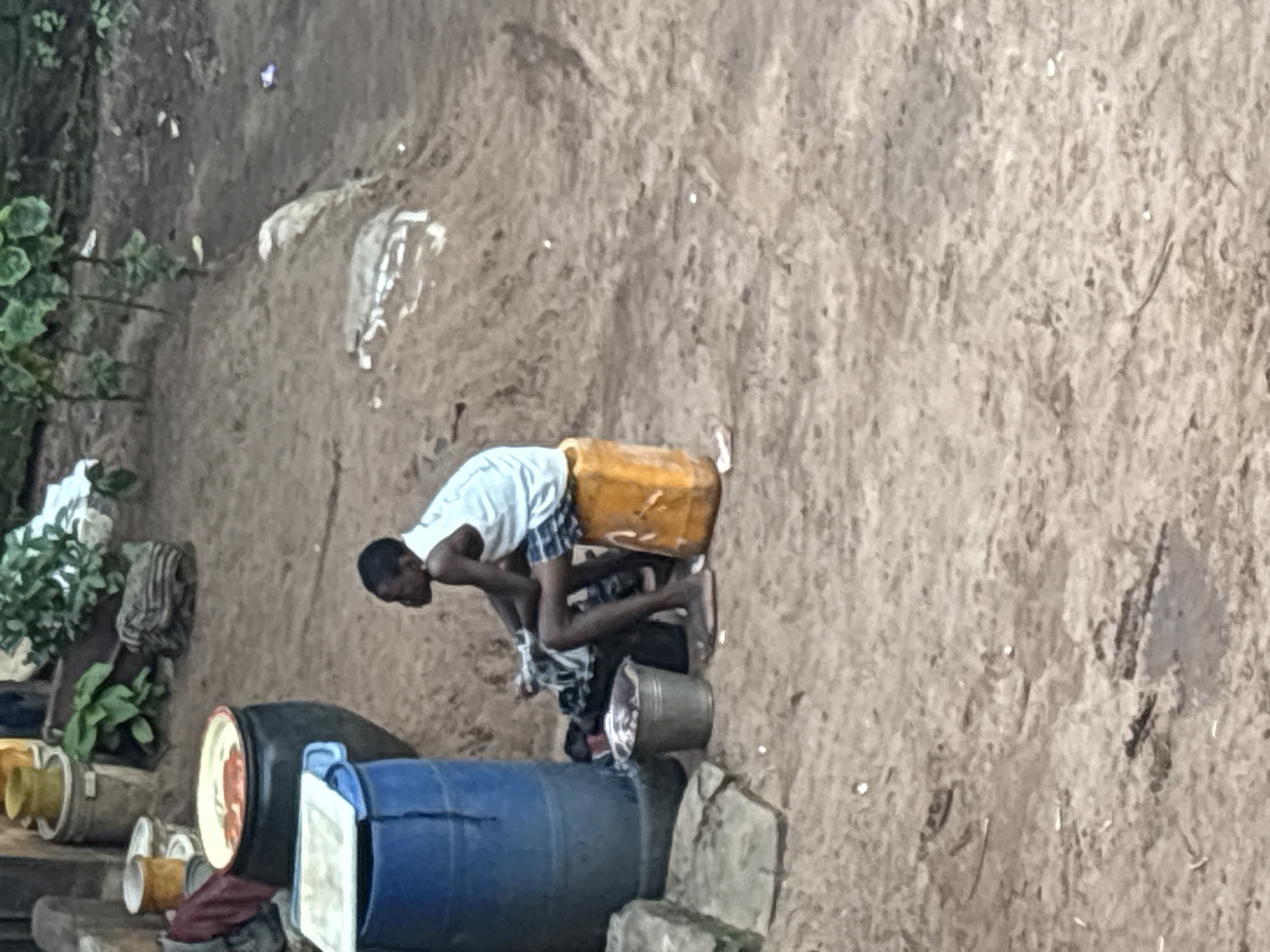A young boy is sitting on a yellow plastic container outdoors on a dirt ground. He is wearing a white tank top and blue and white checkered shorts. To his left is a blue plastic barrel, and in front of him is a silver bucket. Behind him, there are several large black containers. The background shows vegetation and more dirt ground. The scene appears to be in a residential or communal area in Onitsha, Nigeria, likely a backyard or an open space near homes.