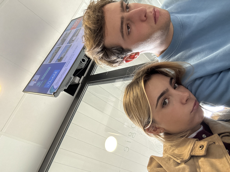 This image captures two young adults in the waiting area of Imagerie Médicale du Sud-Est Lyonnais, located at 10 Rue du Dauphiné, Saint-Priest, France. The female on the left, with blonde hair, rests her head on the right shoulder of the male, who has brown hair. She looks towards the camera with a neutral expression, wearing a tan jacket over a dark inner layer. The male looks down and to his right, away from the camera, wearing a light blue sweatshirt. Above them, a flat-screen monitor displays medical facility information, including "Doctolib," "IMAGERIE MÉDICALE DU SUD-EST LYONNAIS," and the queue number "169." The background shows a white suspended ceiling with integrated lighting and a glass partition reflecting the word "ologie" in reverse, indicating a clinical environment.
