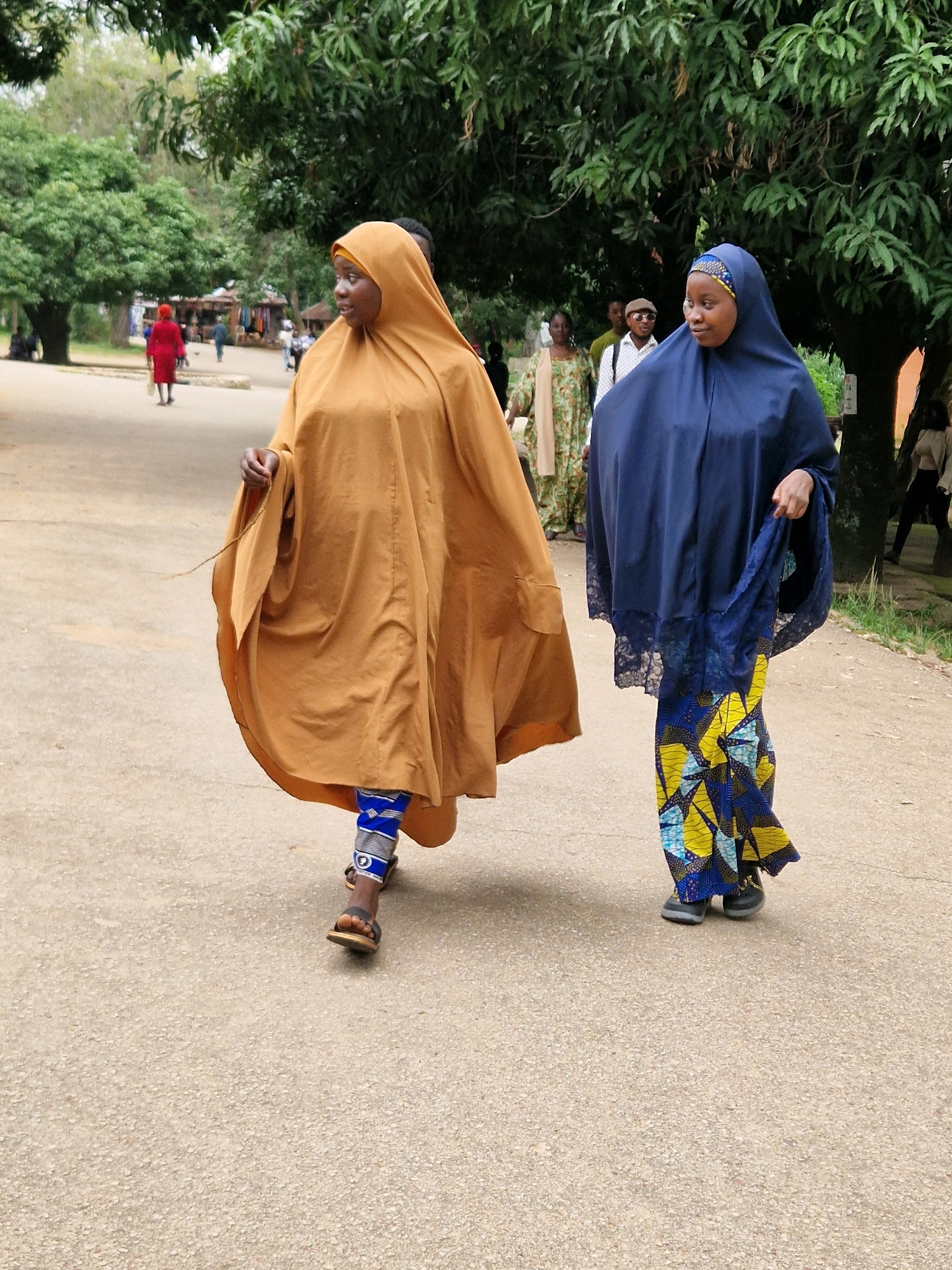 Two women are walking on a paved surface in Jos, Nigeria. The woman on the left is wearing a flowing, tan-colored hijab and dress, with blue and white patterned leggings and brown sandals. The woman on the right is wearing a dark blue hijab and dress with lace trim, and a brightly colored patterned skirt in yellow, blue, and black. She is wearing dark shoes. Both women are facing to the right and appear to be walking together. In the background, there are trees, other people, and market stalls, suggesting a busy outdoor area. The lighting indicates it is daytime, possibly overcast or late afternoon, with soft shadows.