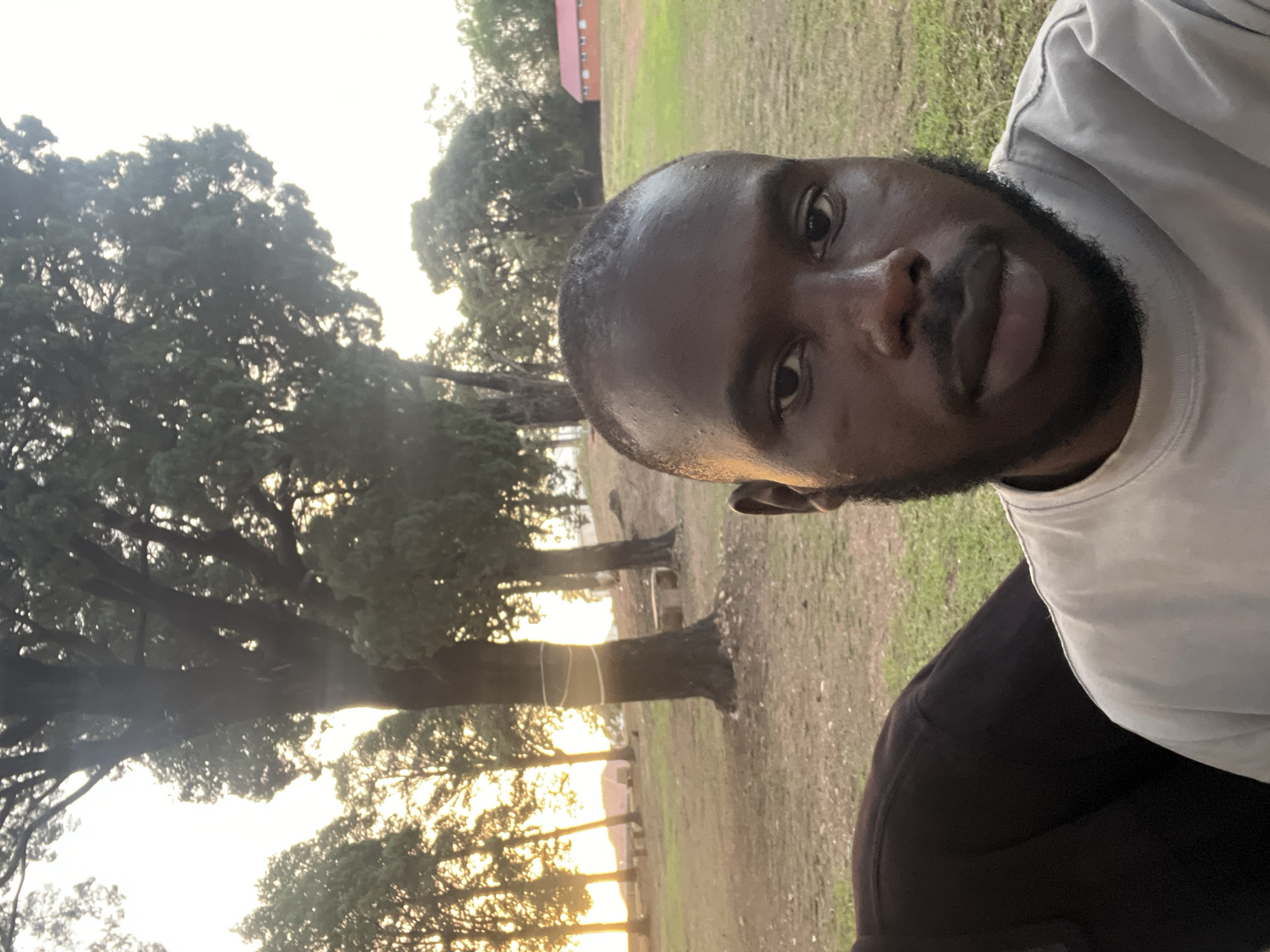 The image is a selfie of a Black man with a beard, wearing a light-colored t-shirt and black pants. He appears to be outdoors, possibly in a park or wooded area, with trees and grass visible in the background. The lighting suggests it is either sunrise or sunset. In the background, there is a building with a red roof. The location appears to be Jos, Nigeria, based on the environment and the man's features.