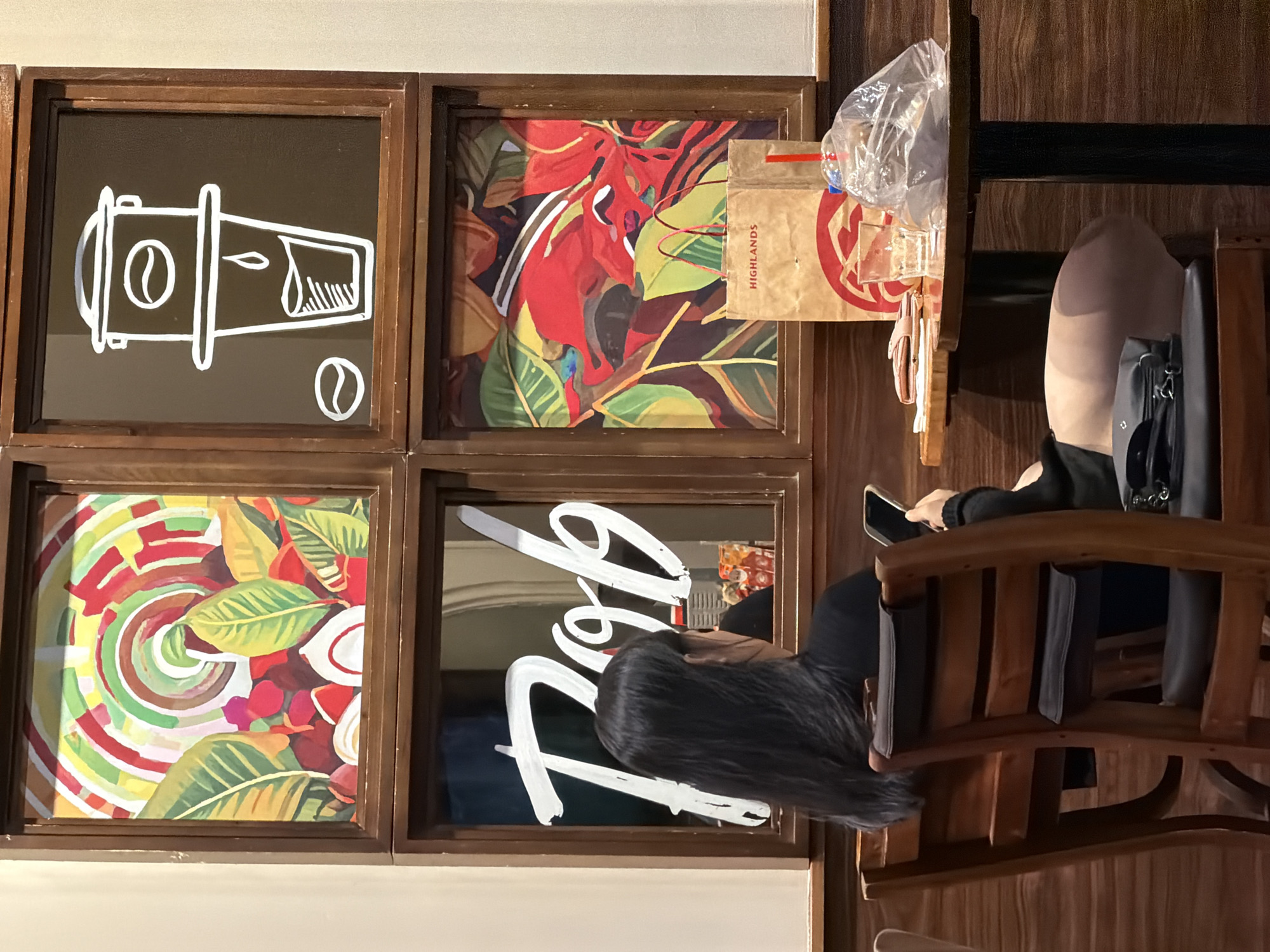 The image depicts an indoor scene, likely a cafe or restaurant, with a young woman seated at a table. She has long, dark hair and is wearing a black top and shorts. She is looking at her phone, which she holds in her right hand. In front of her on the table are a wallet, a glass of water, a paper bag with "HIGHLANDS" printed on it, and a plastic bag containing food or drinks.
The background features a wooden paneling and four framed artworks. The top left artwork is an abstract painting with concentric circles and colorful shapes, interspersed with green leaves. The top right artwork is a minimalist white line drawing of a coffee drip, a coffee bean, and a cup. The bottom left frame is a mirror with white lettering that appears to spell out "Rob." The bottom right artwork is a vibrant painting of red and green foliage. The overall ambiance suggests a casual setting.
The specific location in the city of Nha Trang, Vietnam, is the Highlands Coffee shop located at 75 Tran Phu Street. This is a popular coffee chain in Vietnam, known for its Vietnamese coffee and snacks. The woman is likely a customer enjoying a drink and her phone.