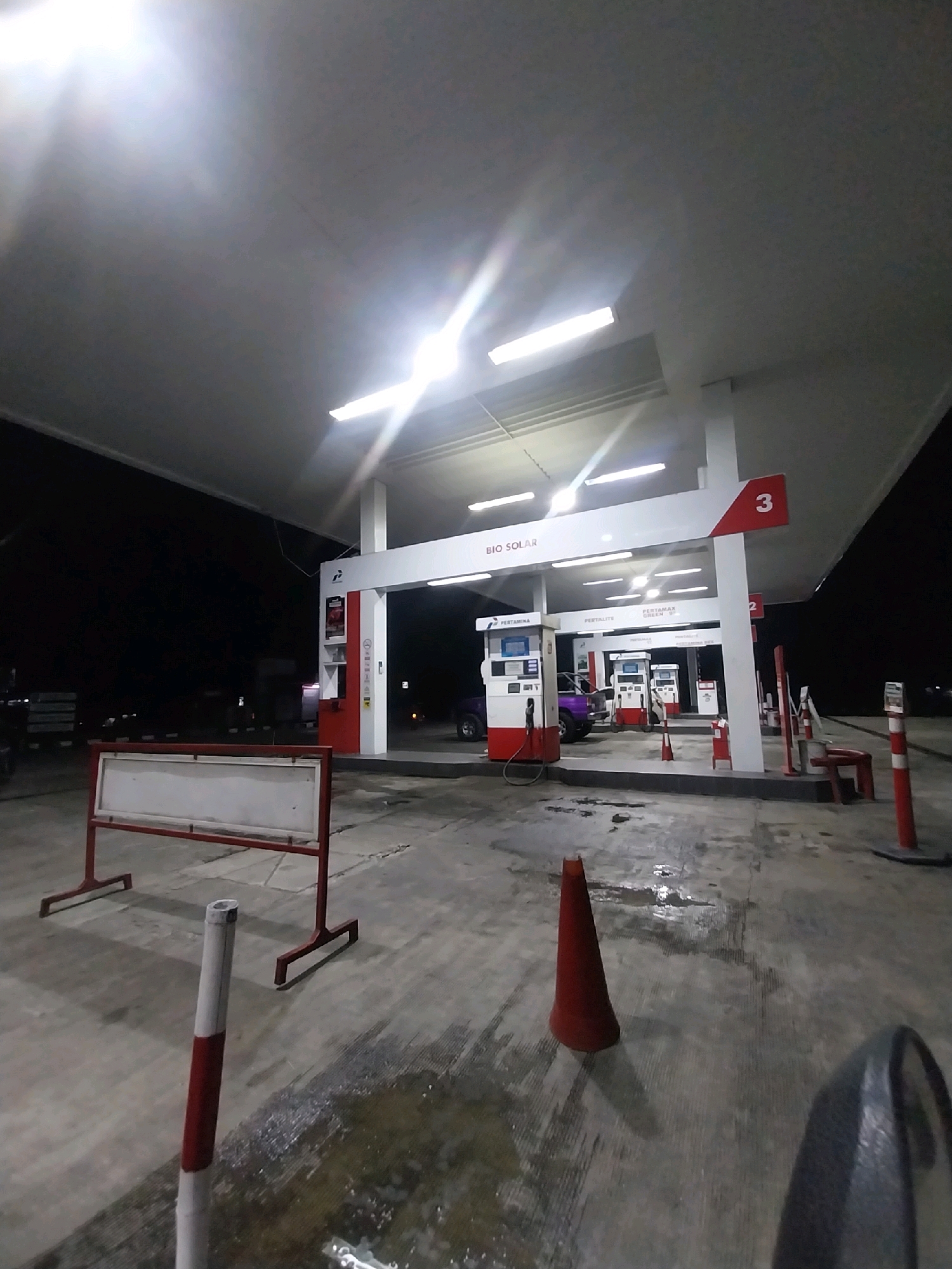 The image depicts a nighttime scene at a Pertamina gas station in Magelang, Indonesia. The foreground features a wet concrete surface with a red traffic cone and a striped bollard. In the midground, there are multiple gas pumps with red and white detailing. Some pumps display screens and branding, including "PERTAMAX GREEN." One pump is labeled with the number "3." A purple vehicle is visible at one of the pumps. A red and white signpost is positioned to the left. The background is dark, suggesting it is late at night. The gas station canopy is illuminated by bright fluorescent lights. The overall environment appears to be an outdoor, open-air location, typical of a service station.