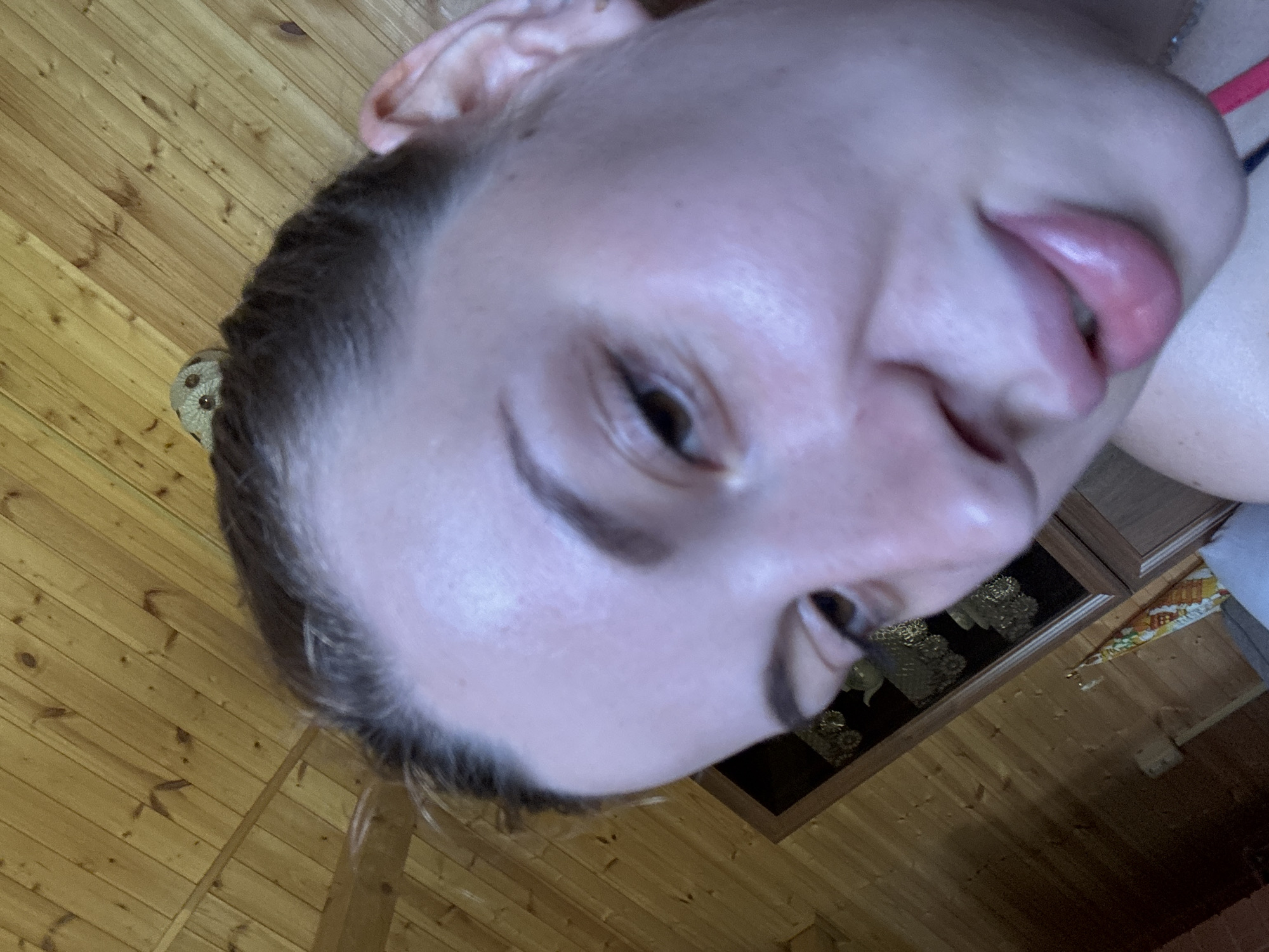 The image shows a close-up, low-angle selfie of a young woman. Her face is partially obscured by the camera's position, with her forehead and the top of her head visible. She has light brown hair pulled back, and her facial features are soft. Her eyes are brown, and her lips are full and pink, with a slight smile. The background consists of a wooden paneled wall and ceiling, consistent with interior architecture. A framed object with golden embellishments is visible on the wall behind her. The overall impression is of a casual, indoor photograph.