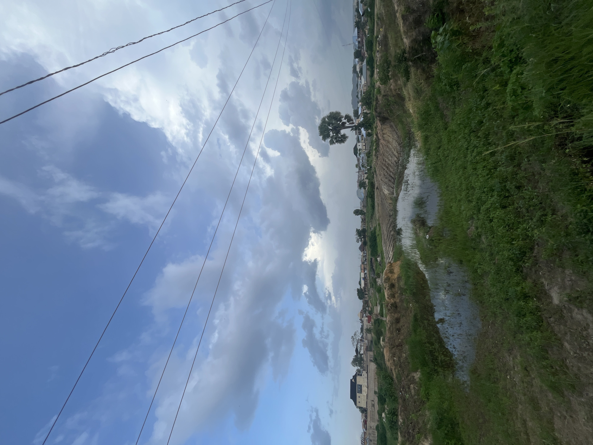 This is a vertical, wide-angle photograph taken in Rafin Par, Nigeria. The image depicts a rural or semi-rural landscape under a cloudy sky. Power lines stretch diagonally across the upper left portion of the frame, silhouetted against the bright sky and scattered clouds.
The scene is divided into sky and land. The sky is a mix of light blue and dramatic, grey clouds, suggesting either an overcast day or approaching rain. A lone palm tree stands out against the sky in the middle ground.
The land shows a contrast between cultivated fields and natural vegetation. To the right, a strip of lush green grass grows along the edge of a body of water, possibly a stream or flooded area, reflecting the sky. Further to the left, terraced fields or cultivated land are visible, with some areas appearing to be recently tilled.
In the distance, a settlement of houses and buildings can be seen nestled along the horizon, indicating human habitation. The architecture appears modest. The overall impression is one of a serene, possibly slightly humid, rural environment. No people or specific activities are clearly depicted, but the presence of the settlement and cultivated land suggests a community living and working in this area. The time of day is not definitively indicated, but the lighting suggests it could be late morning or afternoon. There is no visible text in the image.