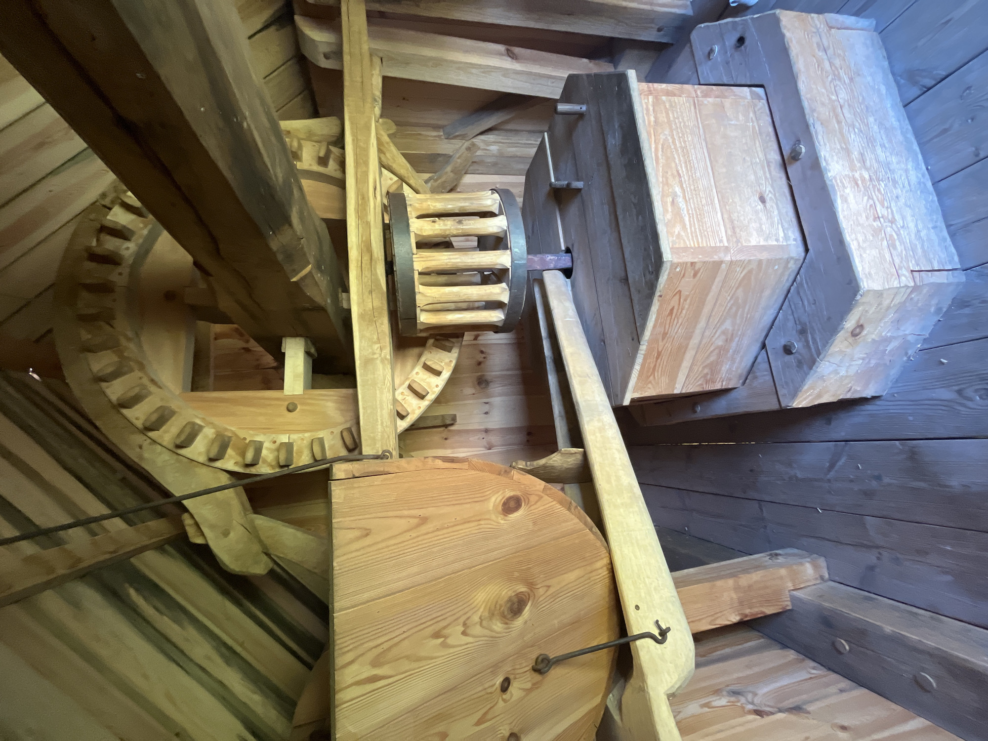 The image shows the interior of a traditional wooden windmill in Kuressaare, Estonia. The scene is dominated by large wooden gears, beams, and a large wooden drum-like structure. The floor is made of wooden planks. In the foreground, there are two wooden boxes, one stacked on top of the other, with a hexagonal shape. The overall construction is rustic and appears to be made of aged wood. There are no people visible in the image. The environment suggests a historical or educational setting, likely a museum or preserved landmark.