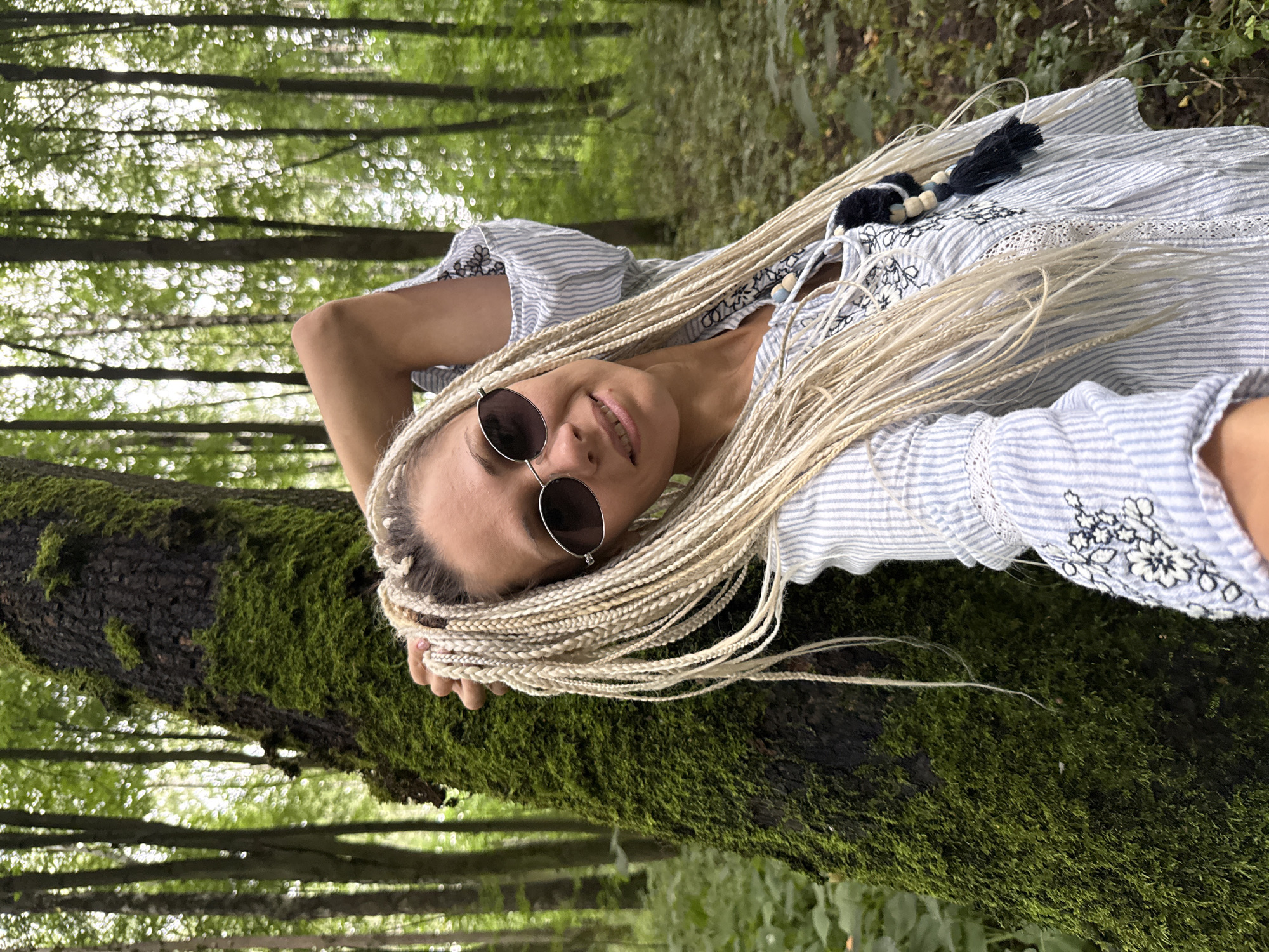 A blonde woman with many thin braids of hair, wearing a light blue and white striped shirt with embroidery and sunglasses, leans against a moss-covered tree in a forest. The background consists of tall trees with green foliage. The location is the village of Rusanovo, Russia.