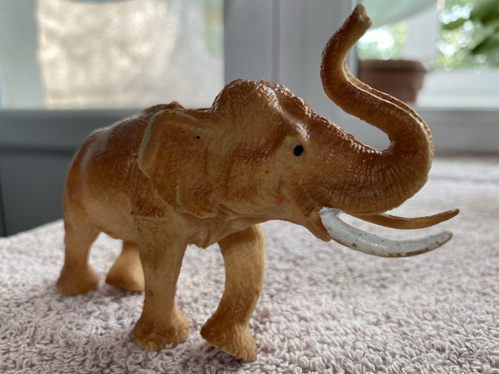An orange plastic toy elephant stands on a pink fuzzy textured surface, possibly a rug or blanket. The elephant is positioned in the center of the frame, facing right, with its trunk raised and curled. Its eyes are small black dots, and it has two tusks. The background is blurred, showing a window with light filtering through and hints of greenery and possibly a potted plant outside. The overall lighting suggests it is daytime. The location is Бескөл ауылдық округі, Kazakhstan.