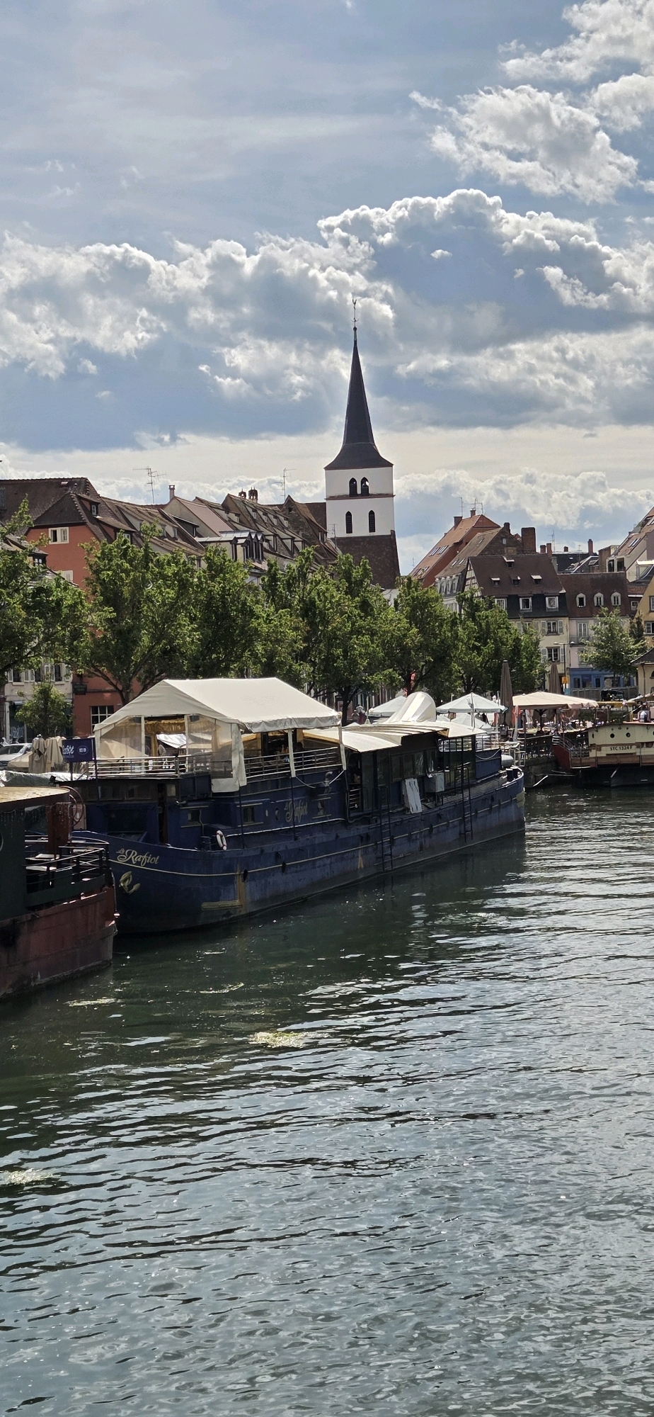 In Strasbourg, France, a calm river flows in the foreground, reflecting the sky and surrounding buildings. Moored along the riverbank is a large, dark blue boat with white awnings covering its deck, appearing to be a restaurant or bar. The boat is named "La Raffiot." Next to it, a portion of a red-brown boat is visible. Behind the boats, a line of traditional Strasbourg buildings with pitched roofs and trees can be seen. Dominating the skyline is a tall church tower with a dark spire and a white section featuring two arched windows. The sky is partly cloudy with dramatic white clouds against a blue background.